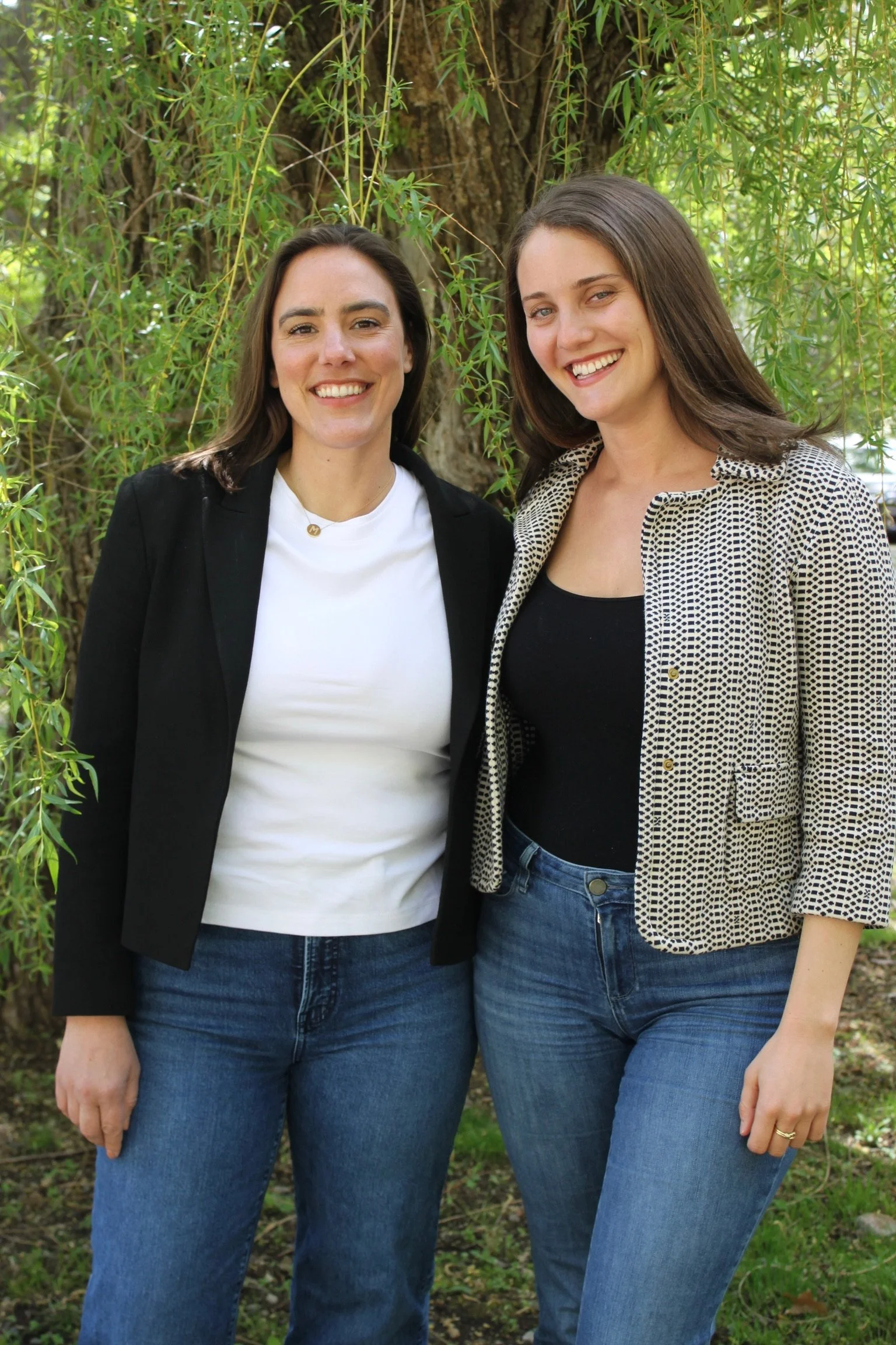 Two women standing outdoors in front of a tree with green leaves, smiling at the camera, wearing casual clothing with jeans.