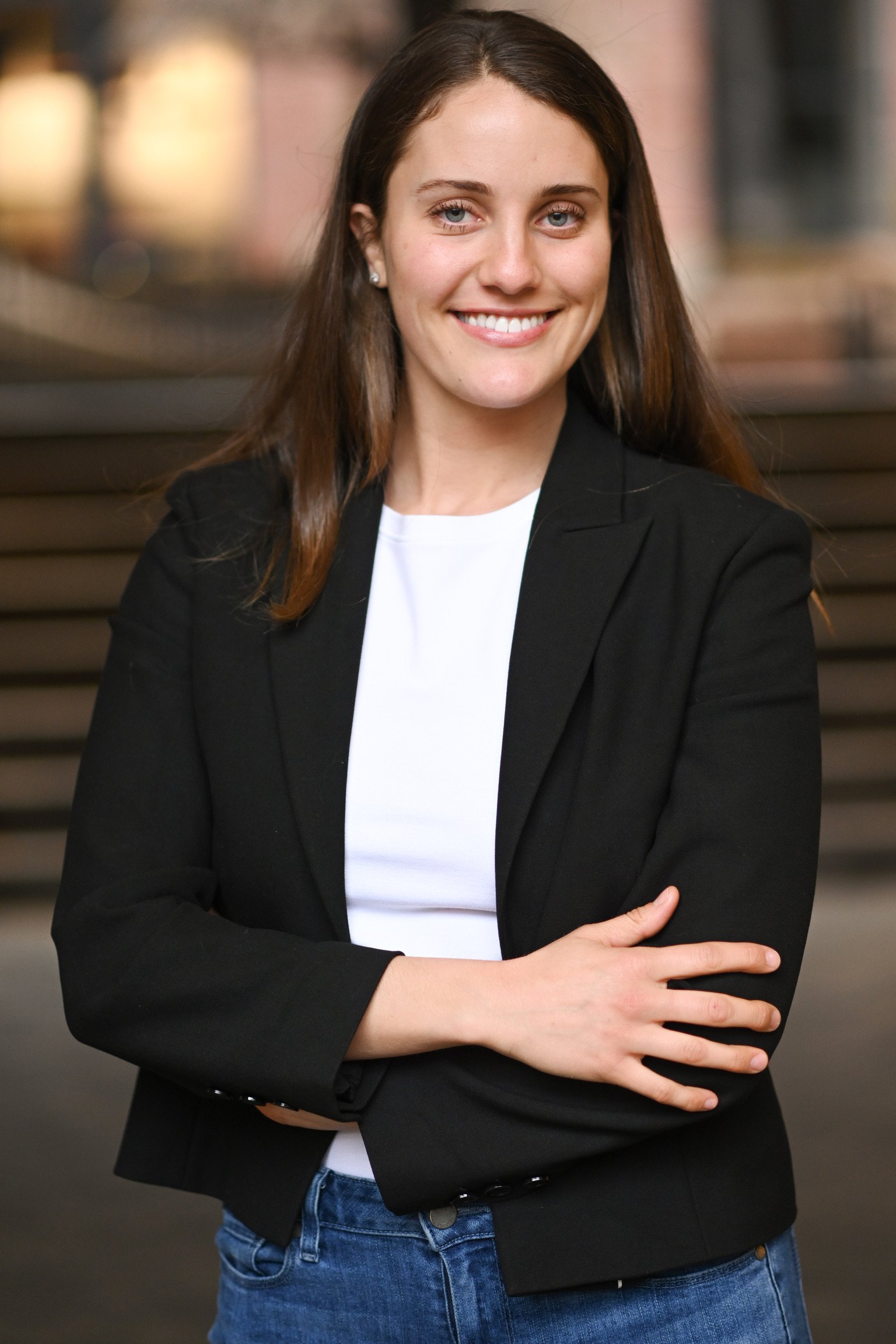 A woman with long brown hair, wearing a black blazer over a white shirt, standing outdoors with arms crossed, smiling at the camera.