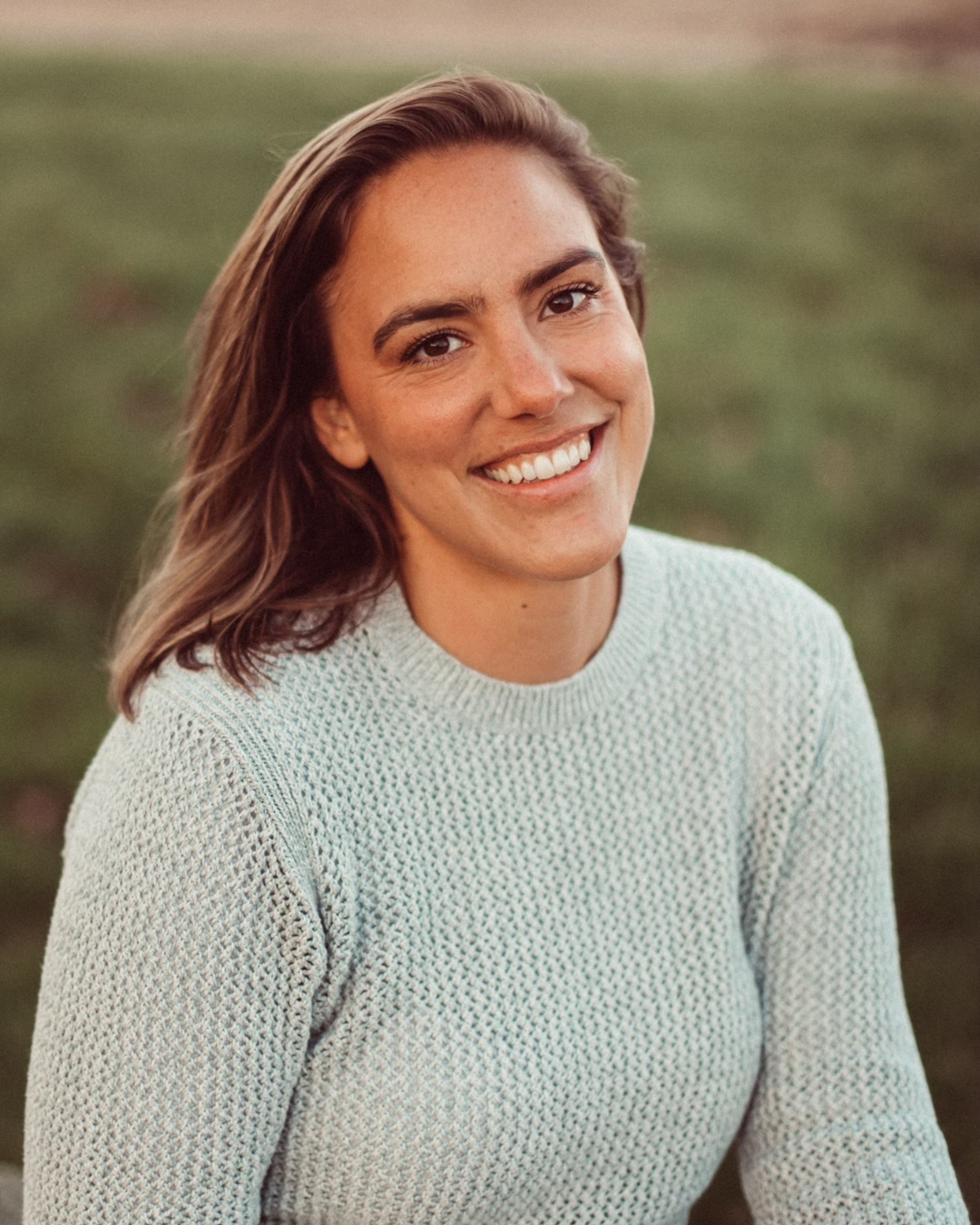 A woman with wavy brown hair and a bright smile, wearing a light-colored knitted sweater, outdoors on a grassy field during sunset.