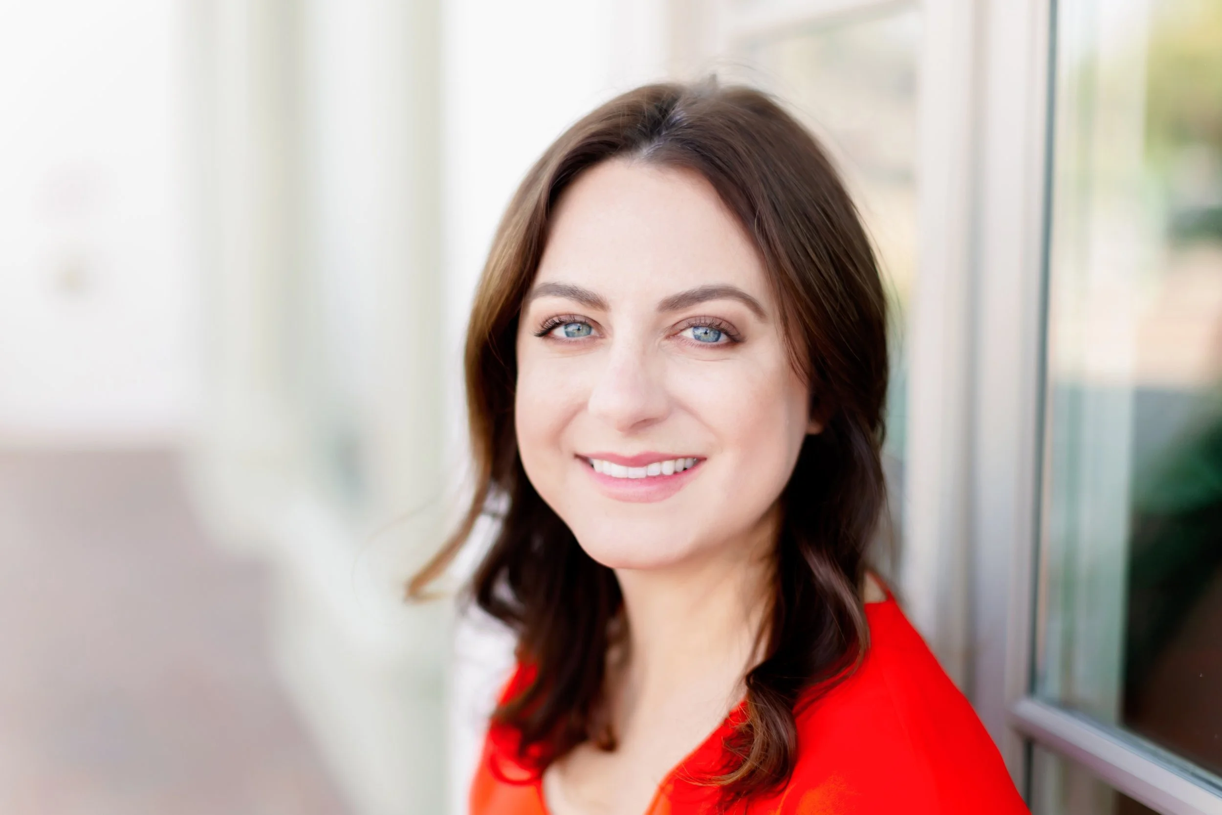 A smiling woman with blue eyes and brown hair wearing a red top, standing outdoors near a window with a blurred background.
