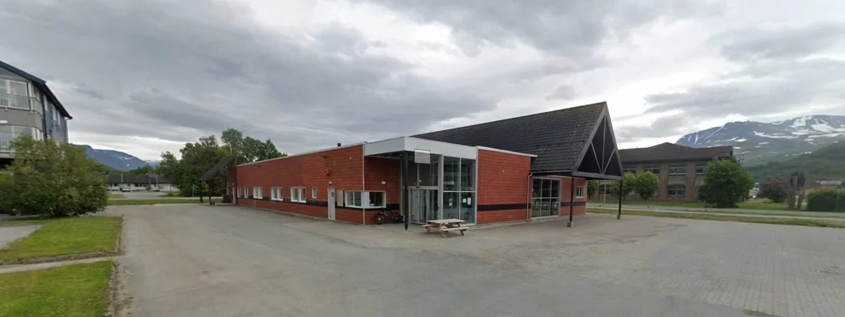 En enkel bygning i rødt murstein med store vinduer, omgitt av en åpen parkeringsplass og grønne områder, med fjell i bakgrunnen.