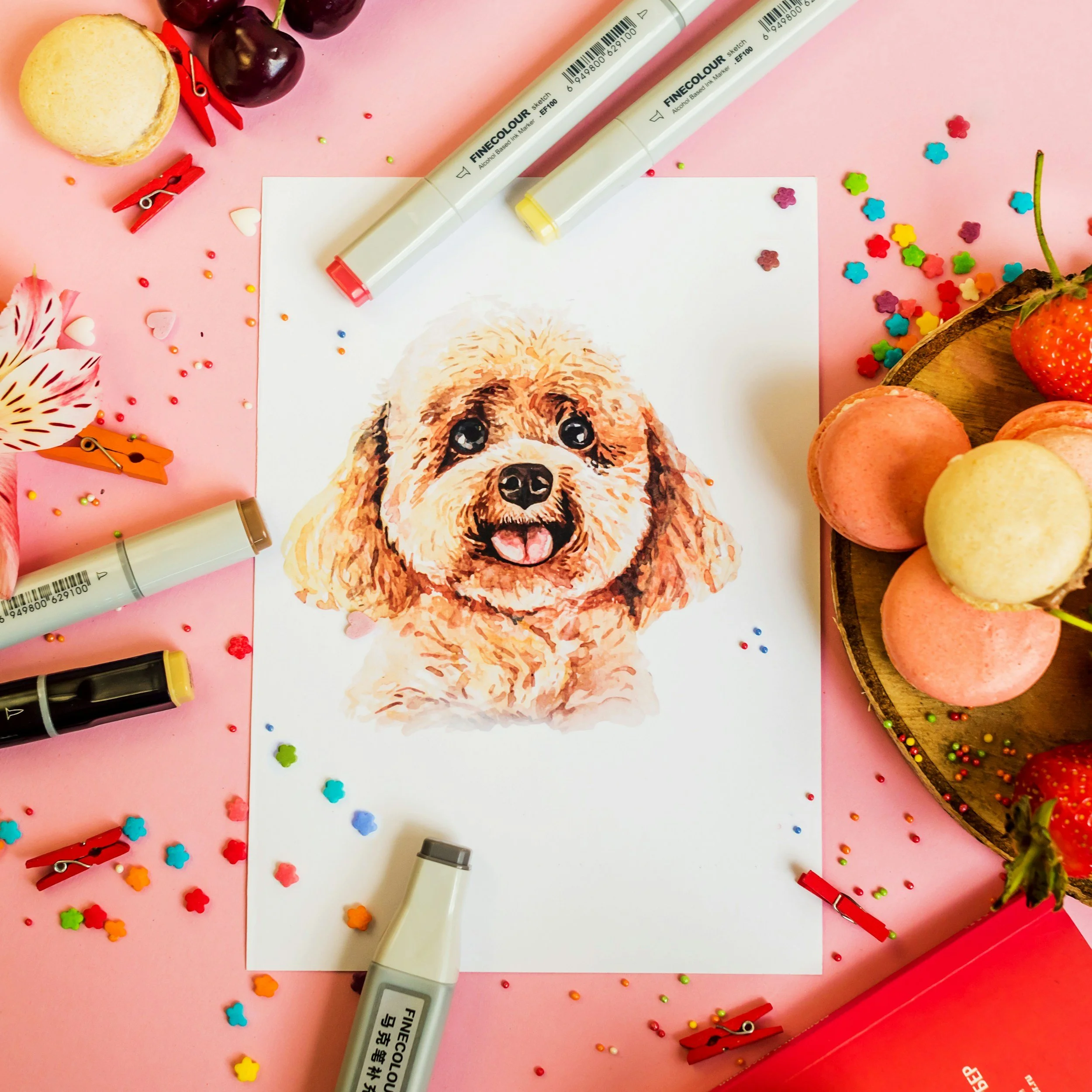 Pink surface with scattered colorful flower-shaped confetti, small round confetti, three white marker pens, a drawing of a dog, and various decorative items including cherries, a strawberry, macarons, and clothespins.