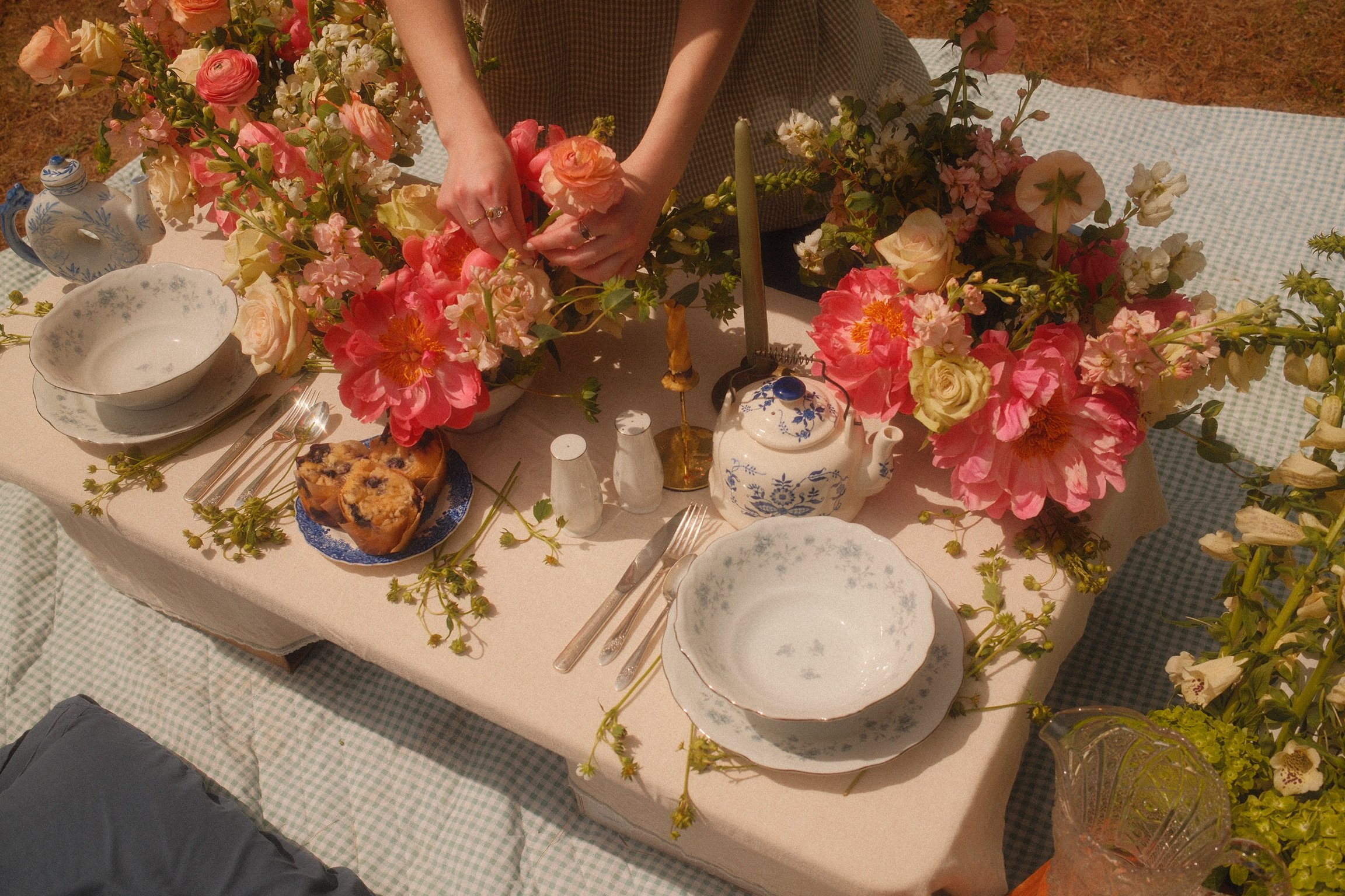 A person arranging pink and cream flowers on a white tablecloth covered table set for a meal, with plates, cutlery, and vintage teapots.