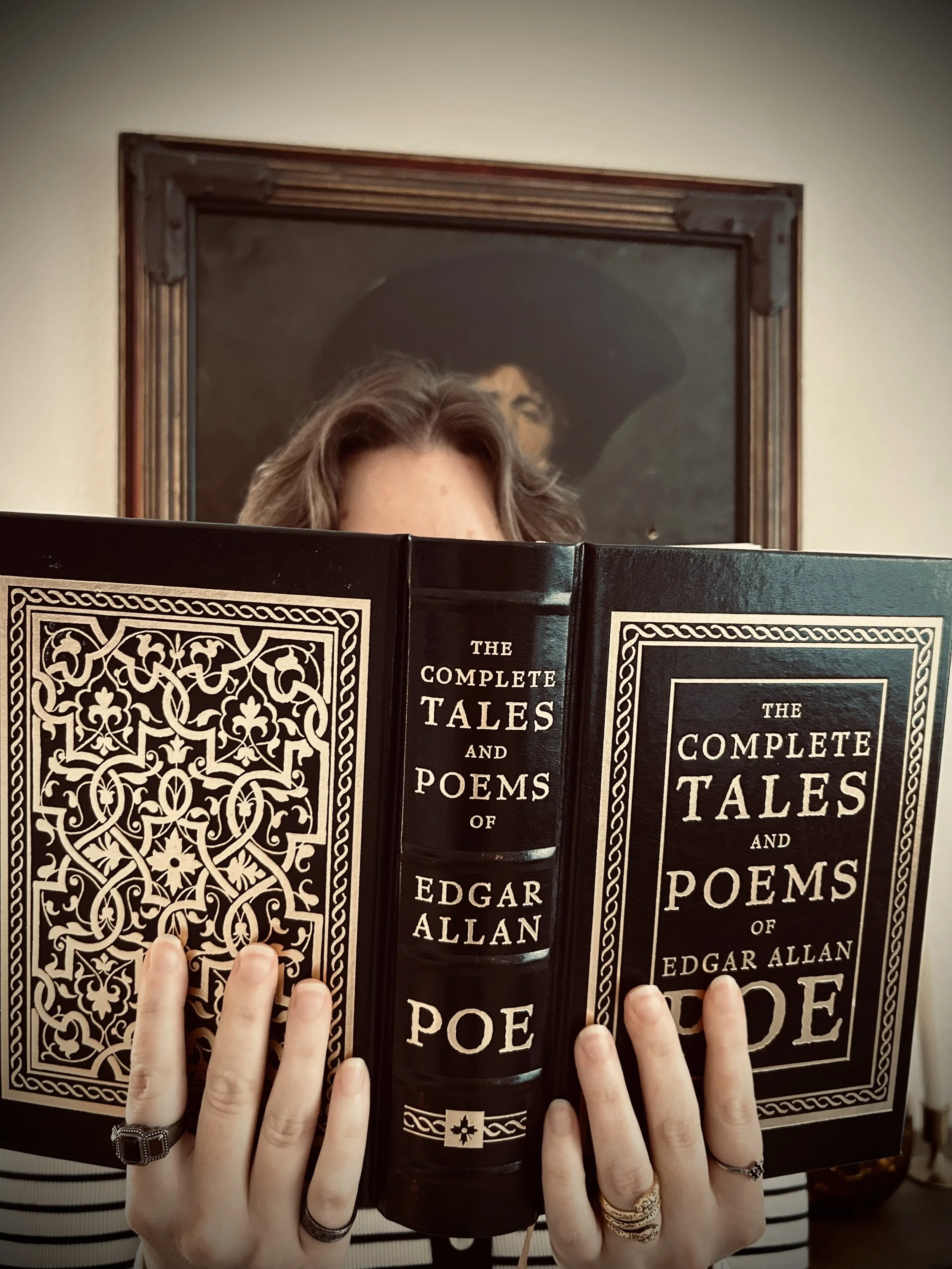 Person holding a large copy of 'The Complete Tales and Poems of Edgar Allan Poe' in front of their face, with a framed portrait of a man in a dark hat hanging on the wall behind them.