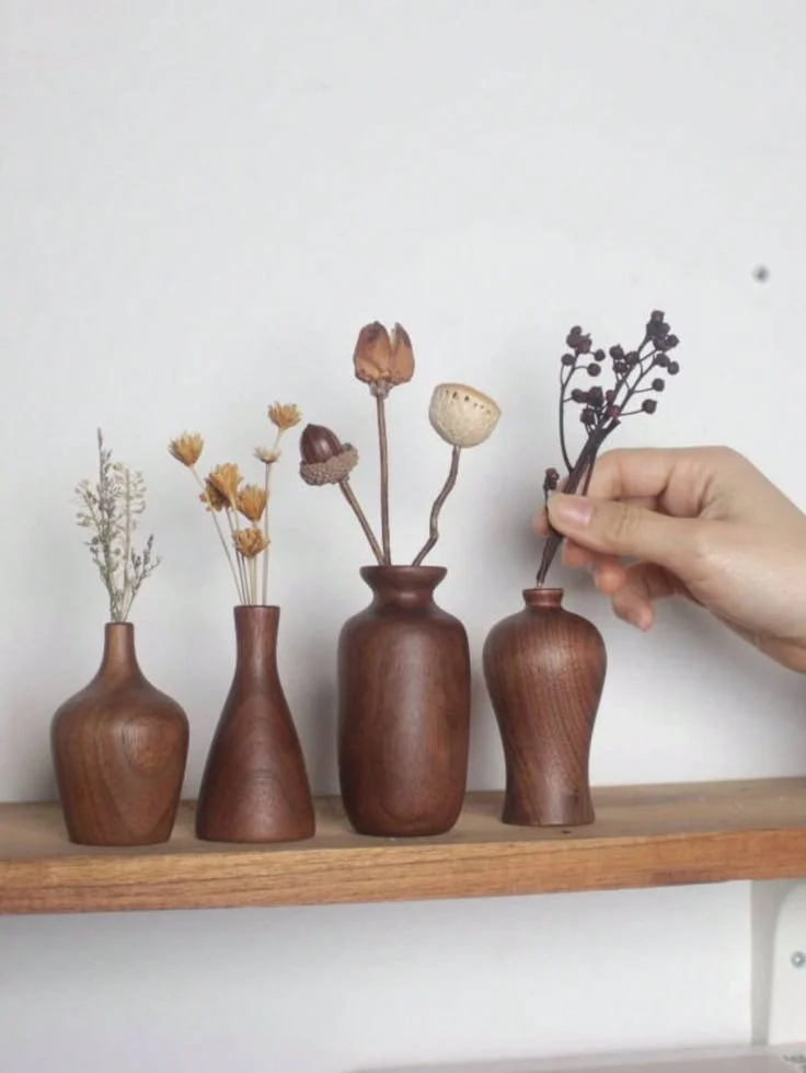 Four small wooden vases with dried flowers and branches are arranged on a wooden shelf, with a person's hand placing or adjusting a branch in the first vase on the right.