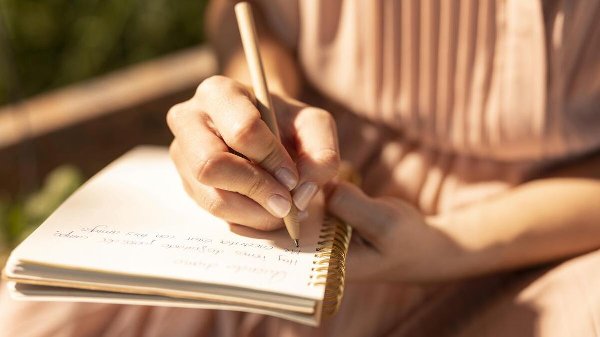 A person writing in a spiral notebook outdoors during daytime, with a blurred background.