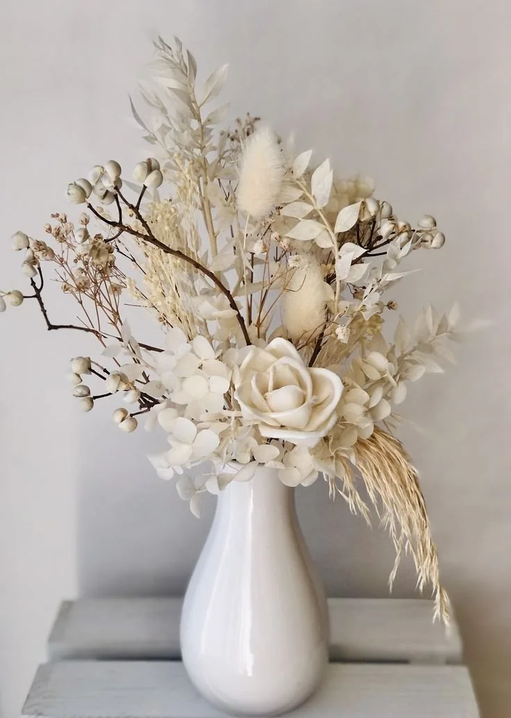 White ceramic vase with dried white and cream-colored flowers and foliage on a light wooden surface