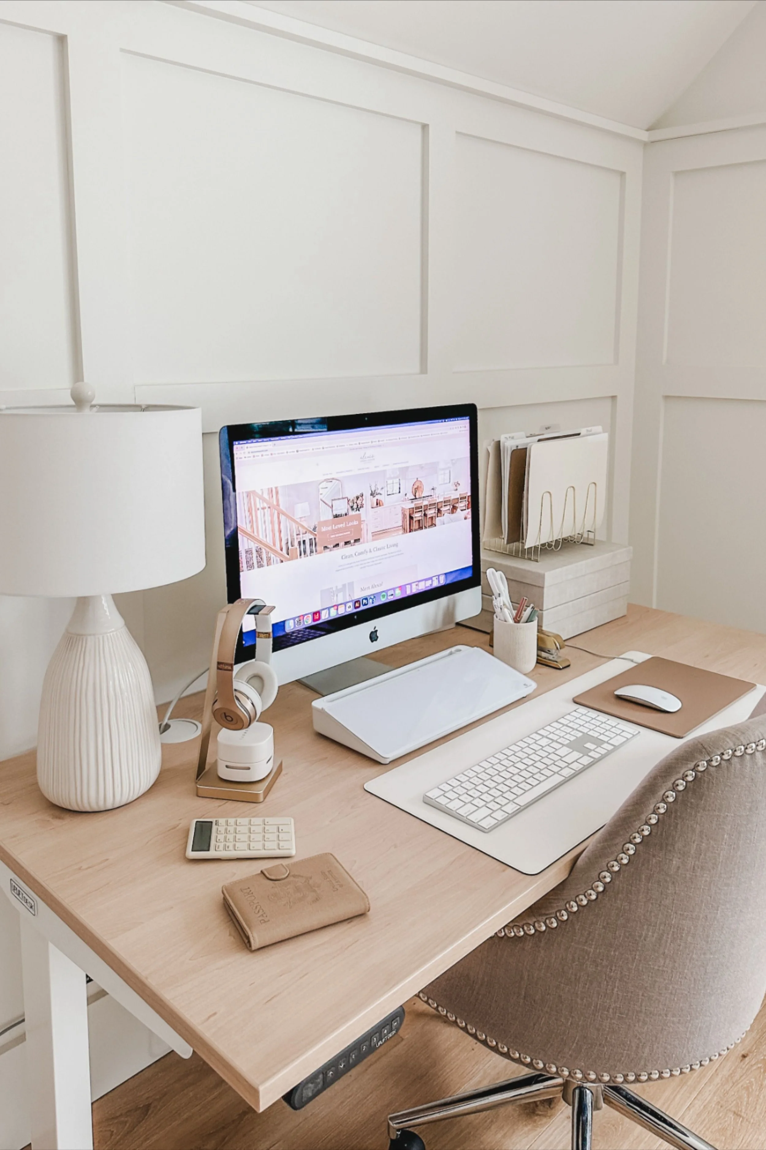 A tidy workspace with a computer monitor, keyboard, mouse, and various office supplies on a light wood desk. A beige, upholstered office chair with pearl embellishments on the arms. A lamp, headphones, calculator, notebooks, and file organizers are also on the desk. The background features white paneled walls.