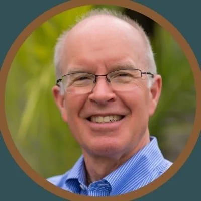 Portrait of a smiling middle-aged man with glasses and a blue striped shirt, outdoors with greenery in the background.