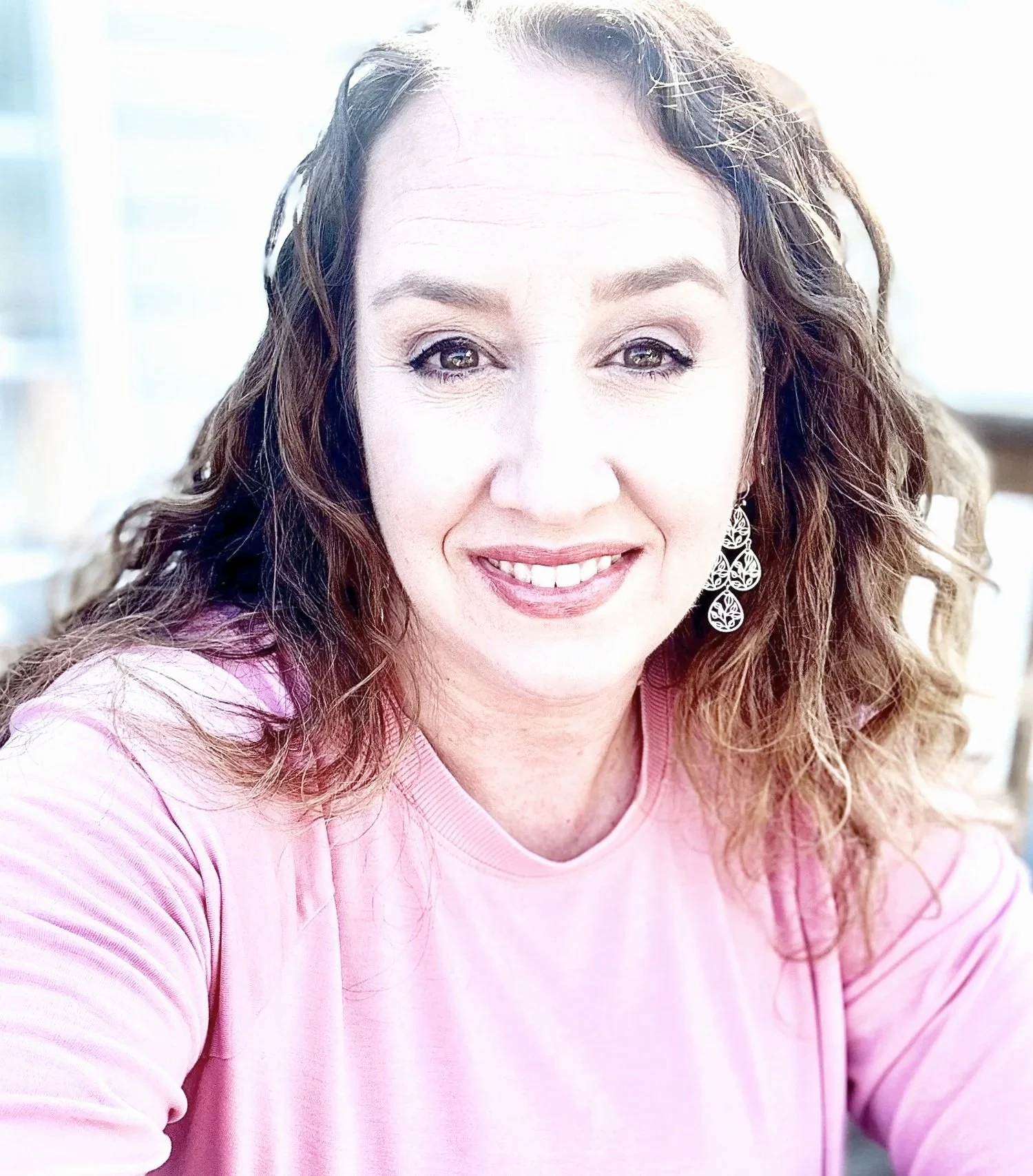 A woman with curly brown hair and earrings smiling at the camera, wearing a pink shirt, in a bright setting.