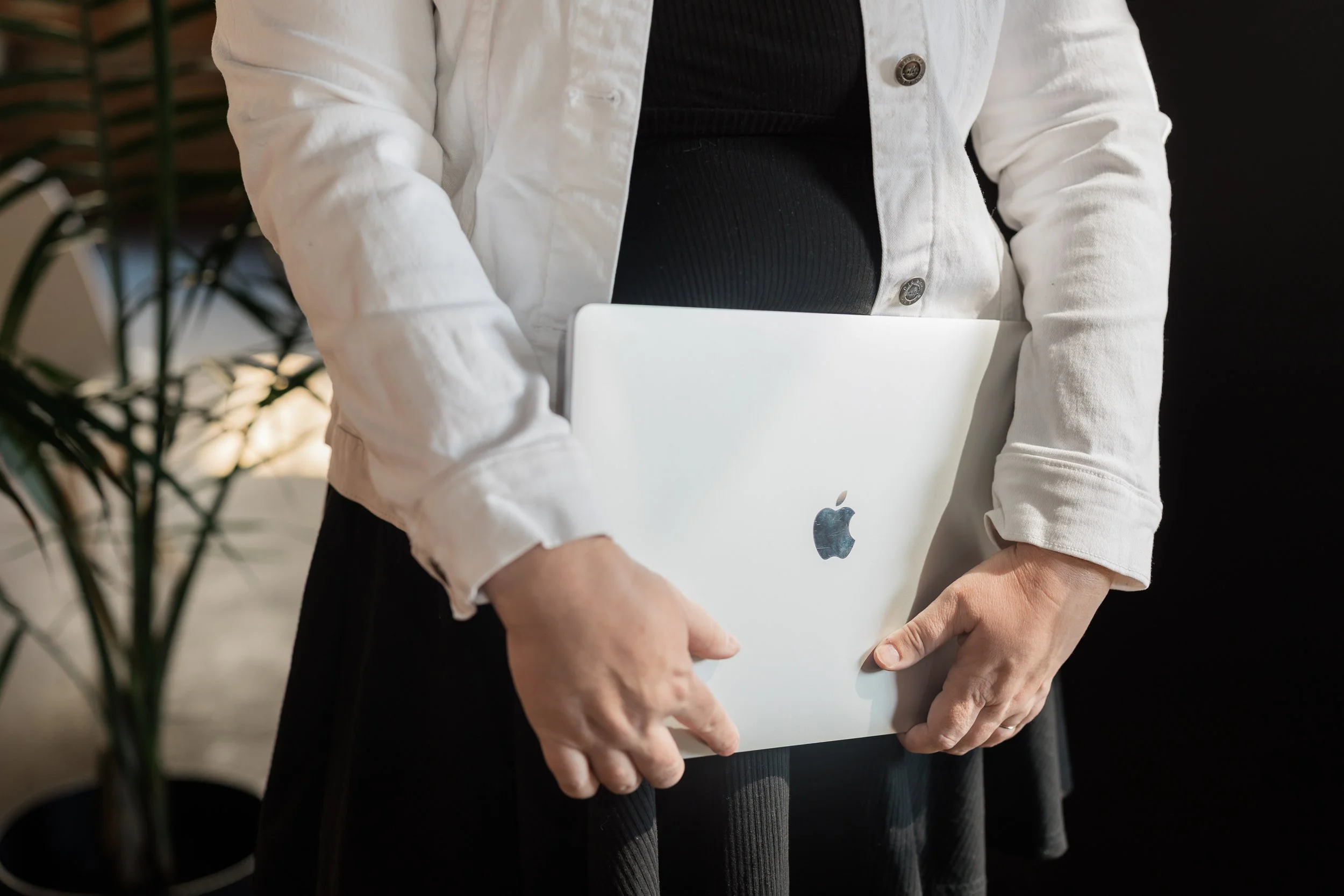 Person holding a closed silver laptop with Apple logo, dressed in a white jacket and black shirt, standing indoors.