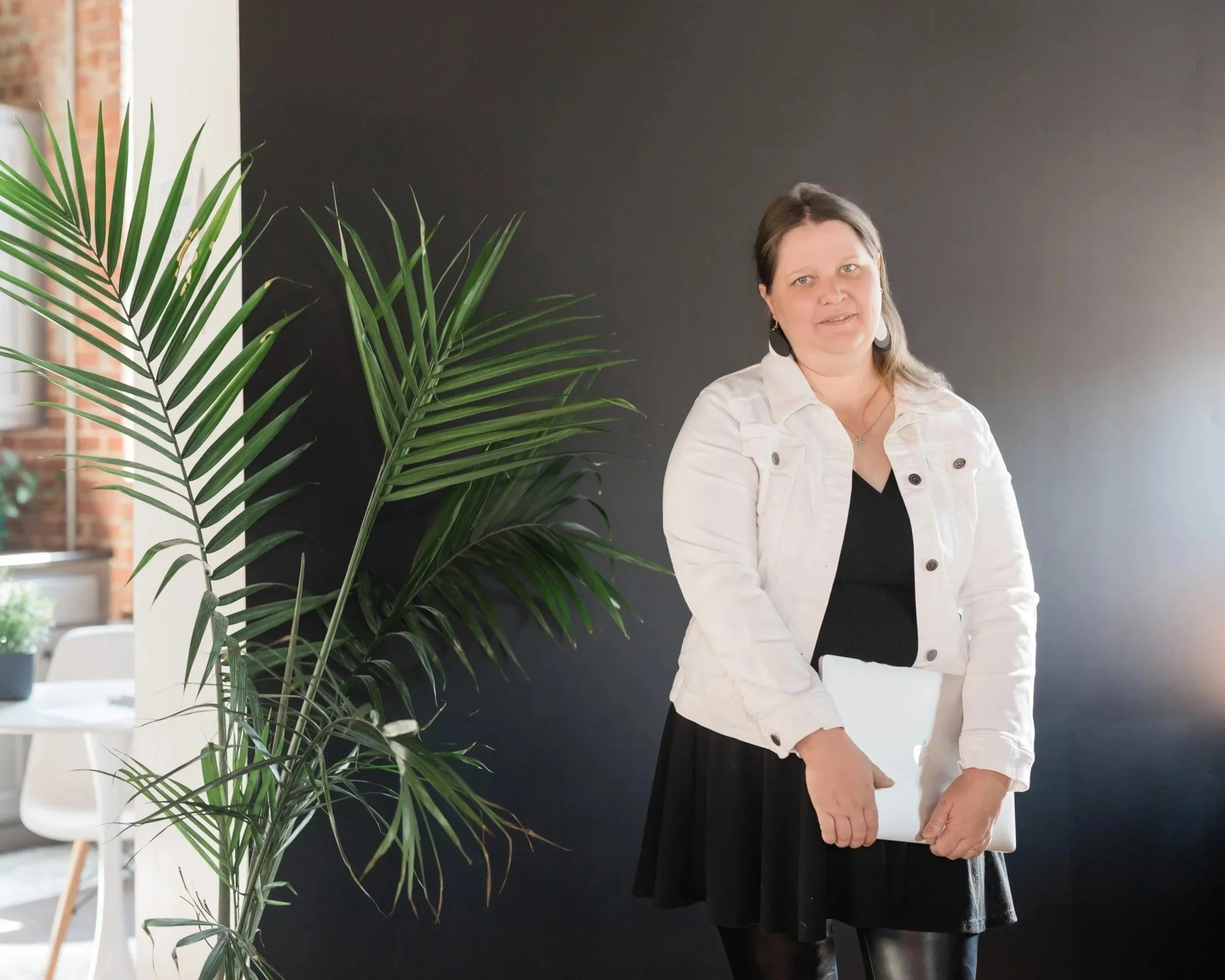 A woman holding a white tablet or notebook in front of her, standing beside a tall green potted plant indoors, with a dark wall behind her and a brick wall partly visible in the background.