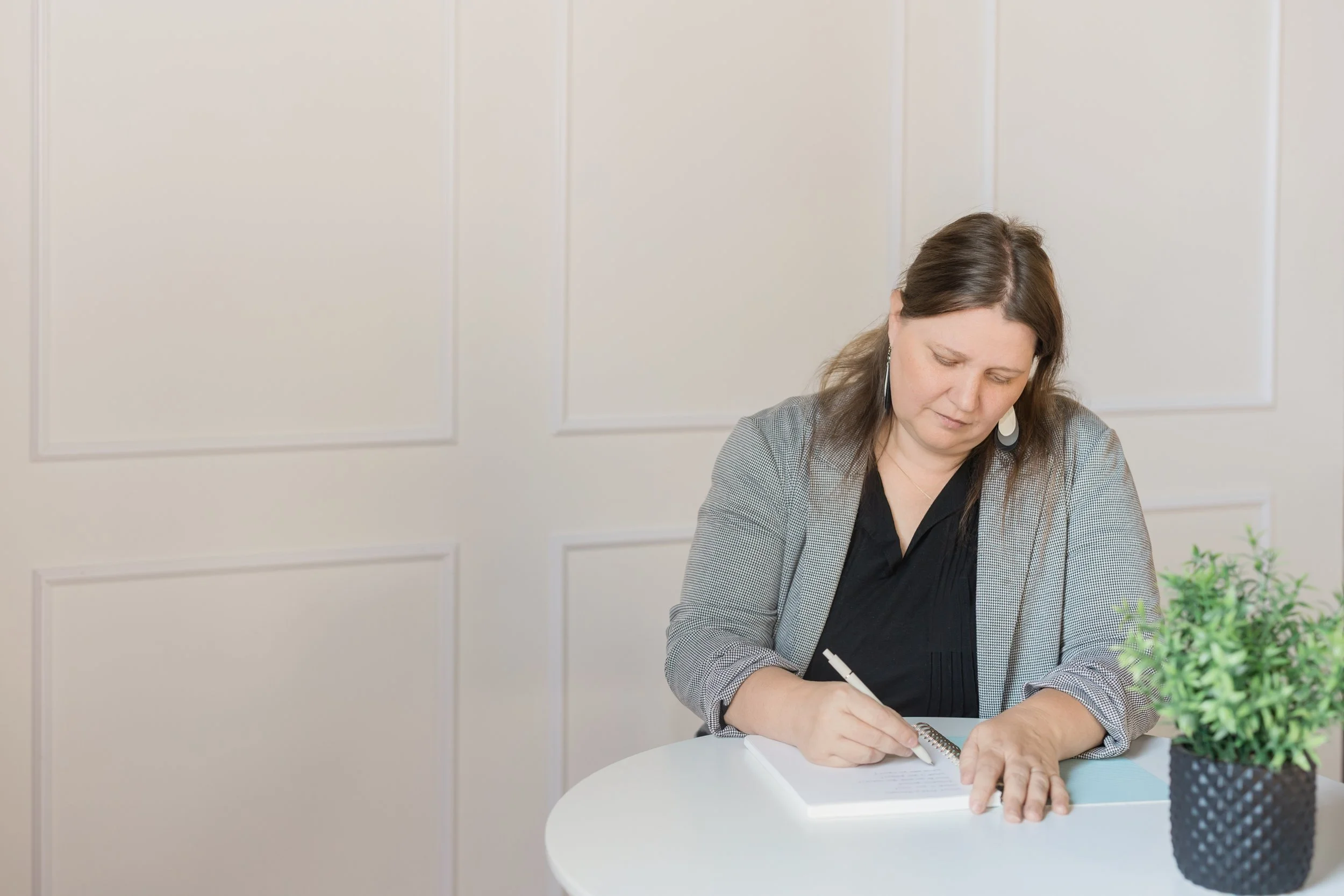 A woman sitting at a white table, writing in a notebook with a pen, against a plain white wall background.