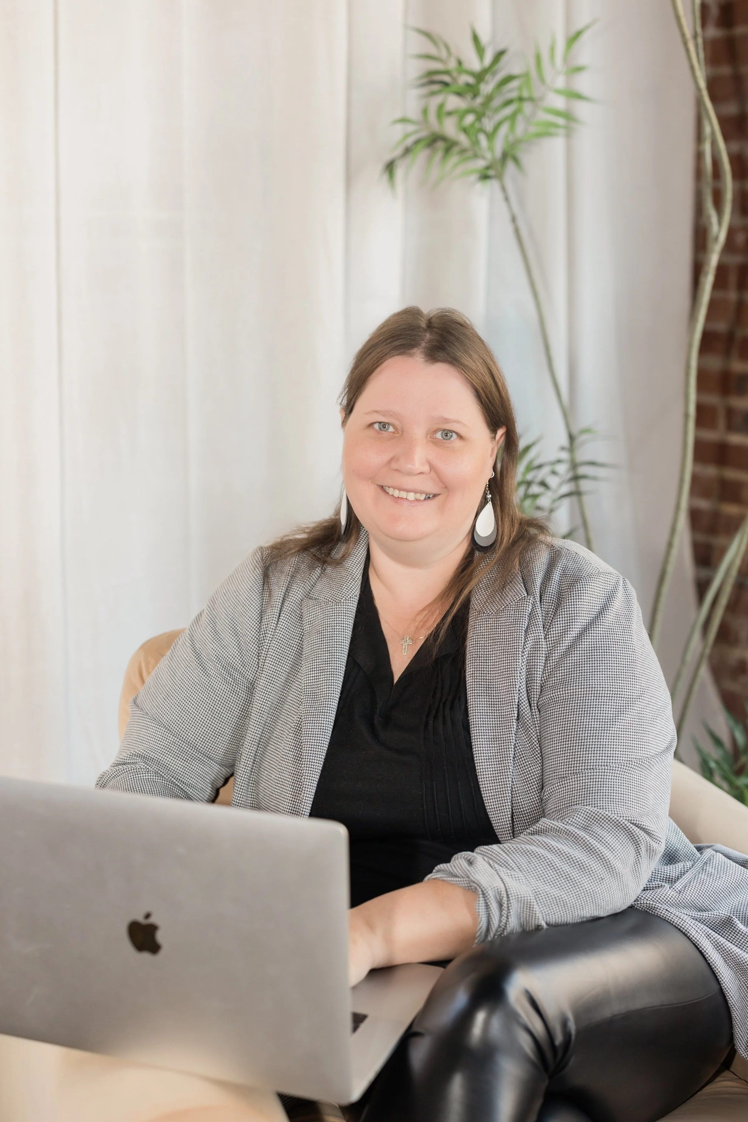 A woman with brown hair and earrings sitting at a desk, working on her laptop, with a plant and a brick wall in the background.