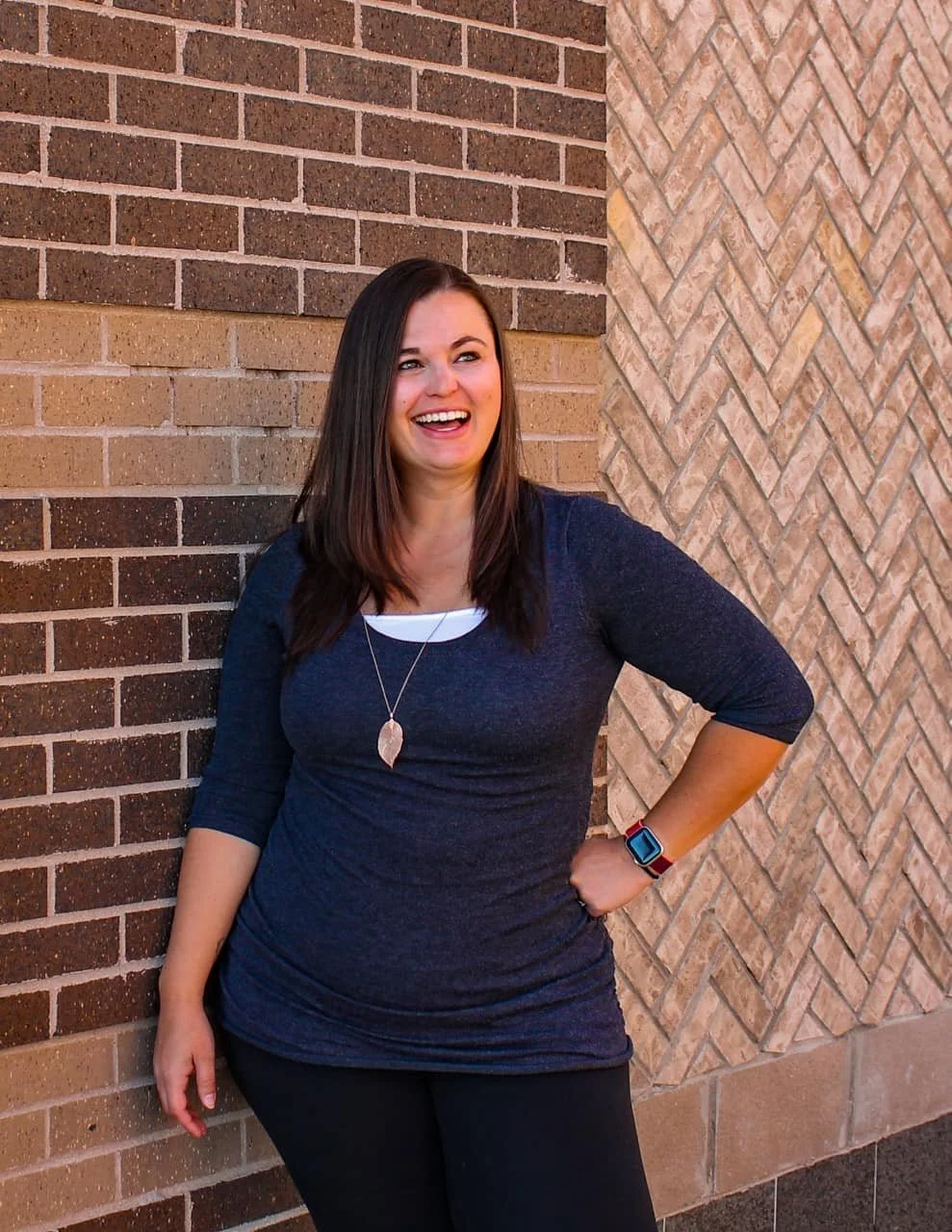 A woman with dark hair, wearing a dark blue long-sleeve shirt and black pants, is standing against a brick wall, smiling and looking to her right. She has a necklace with a leaf pendant and a smartwatch with a red band on her left wrist.