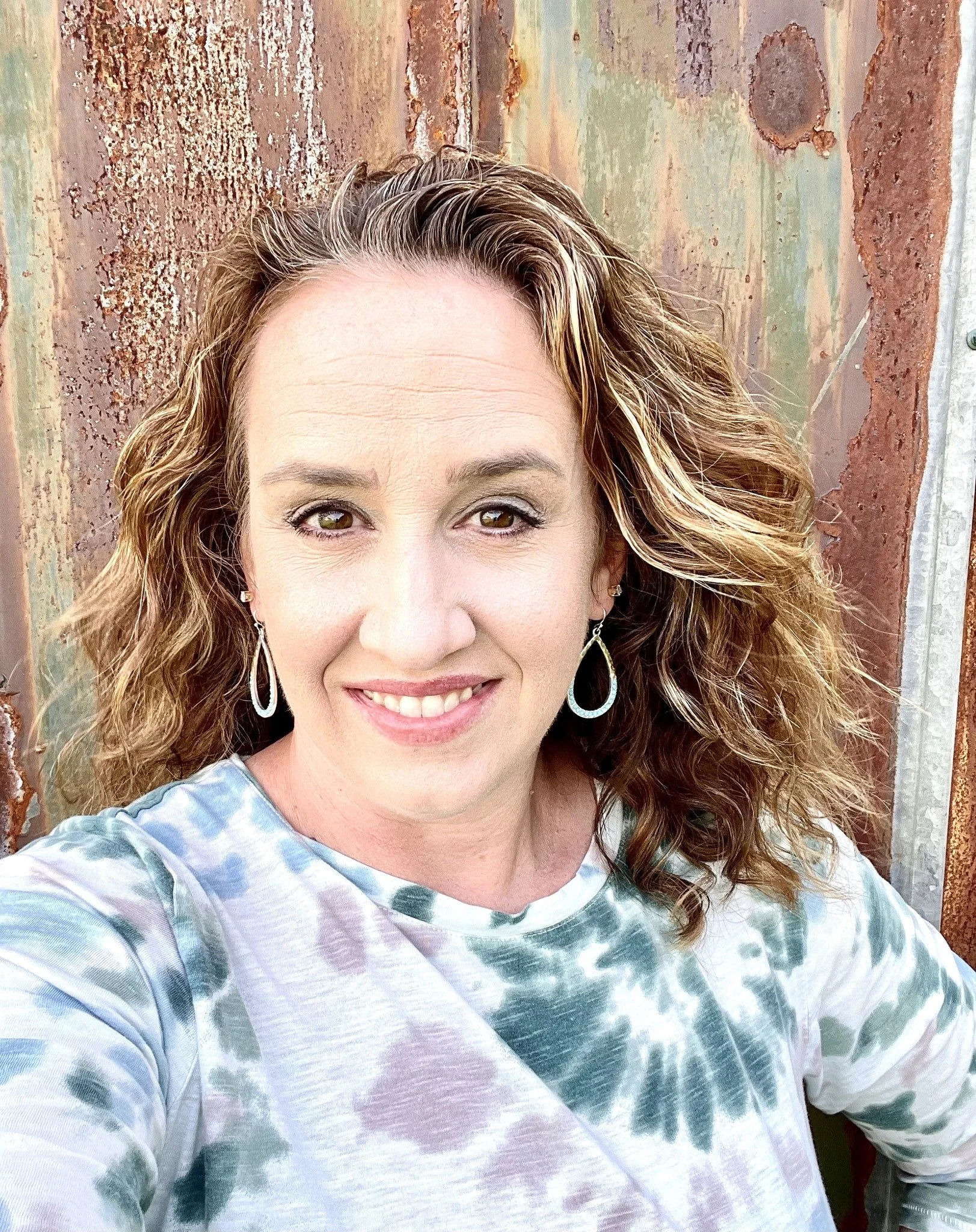 Close-up of a woman with curly auburn hair, wearing earrings, smiling slightly, in front of a rusty metal wall with peeling paint.