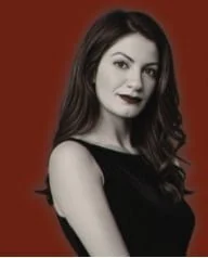 A woman with long dark hair and dark lipstick, wearing a black sleeveless top, posing against a reddish background.