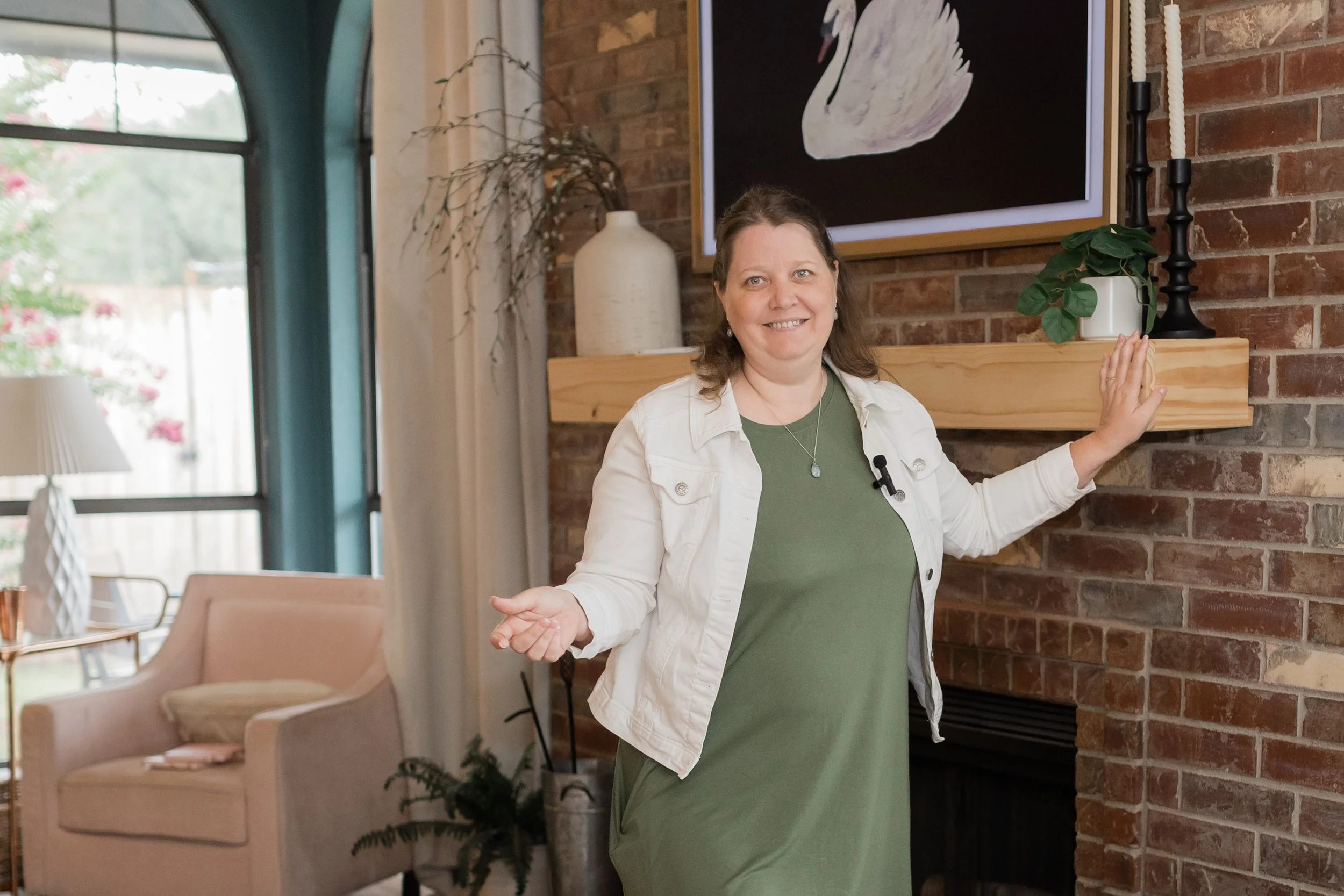 Woman standing in a room with a brick wall, a large framed swan art piece on the wall, and decorative items on a wooden shelf. She is smiling, wearing a green dress and a white jacket, with her right hand resting on the wall and her left hand raised slightly.