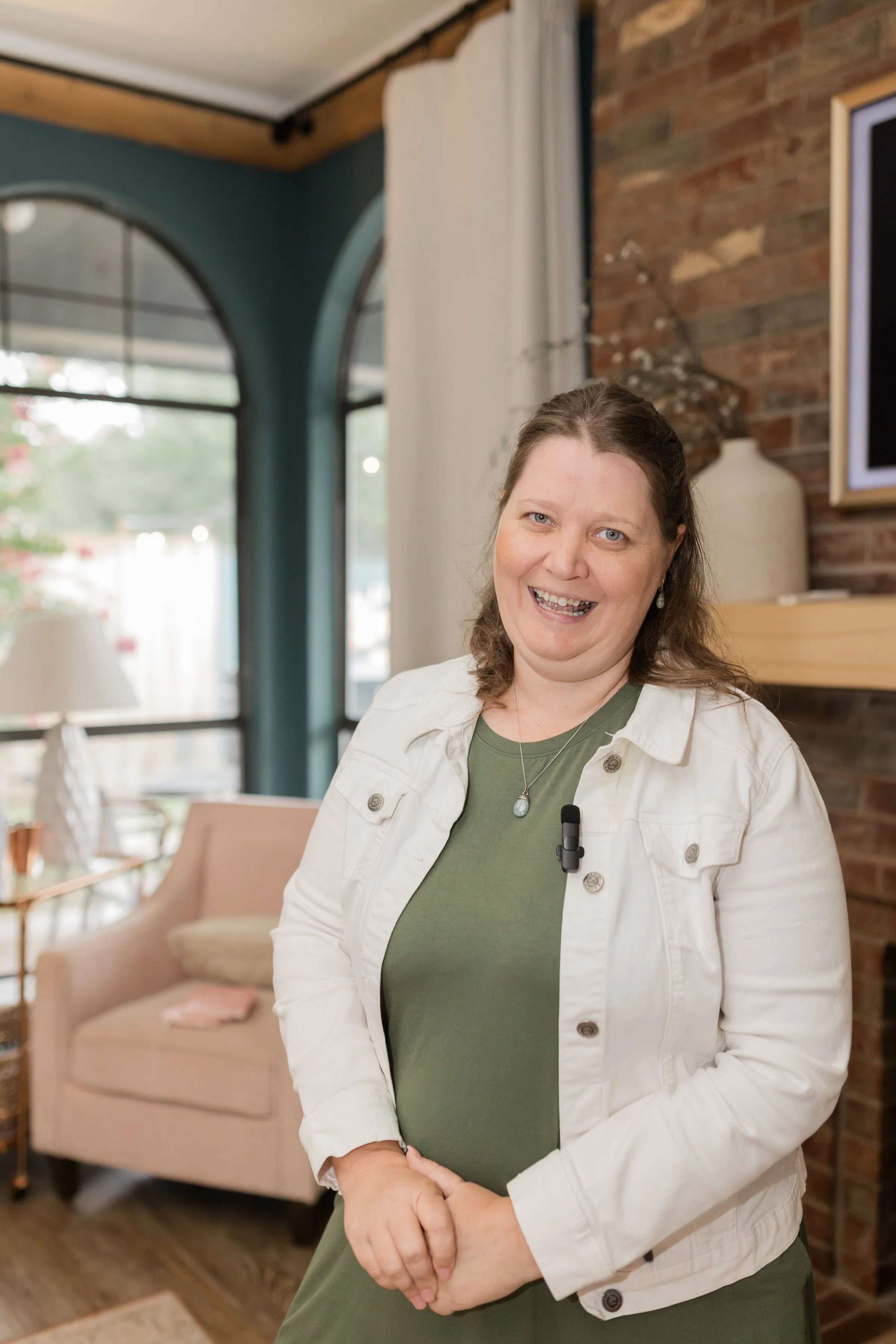A woman with brown hair smiling, wearing a white jacket over a green shirt, standing in a cozy indoor space with a brick wall, a window, and furniture in the background.
