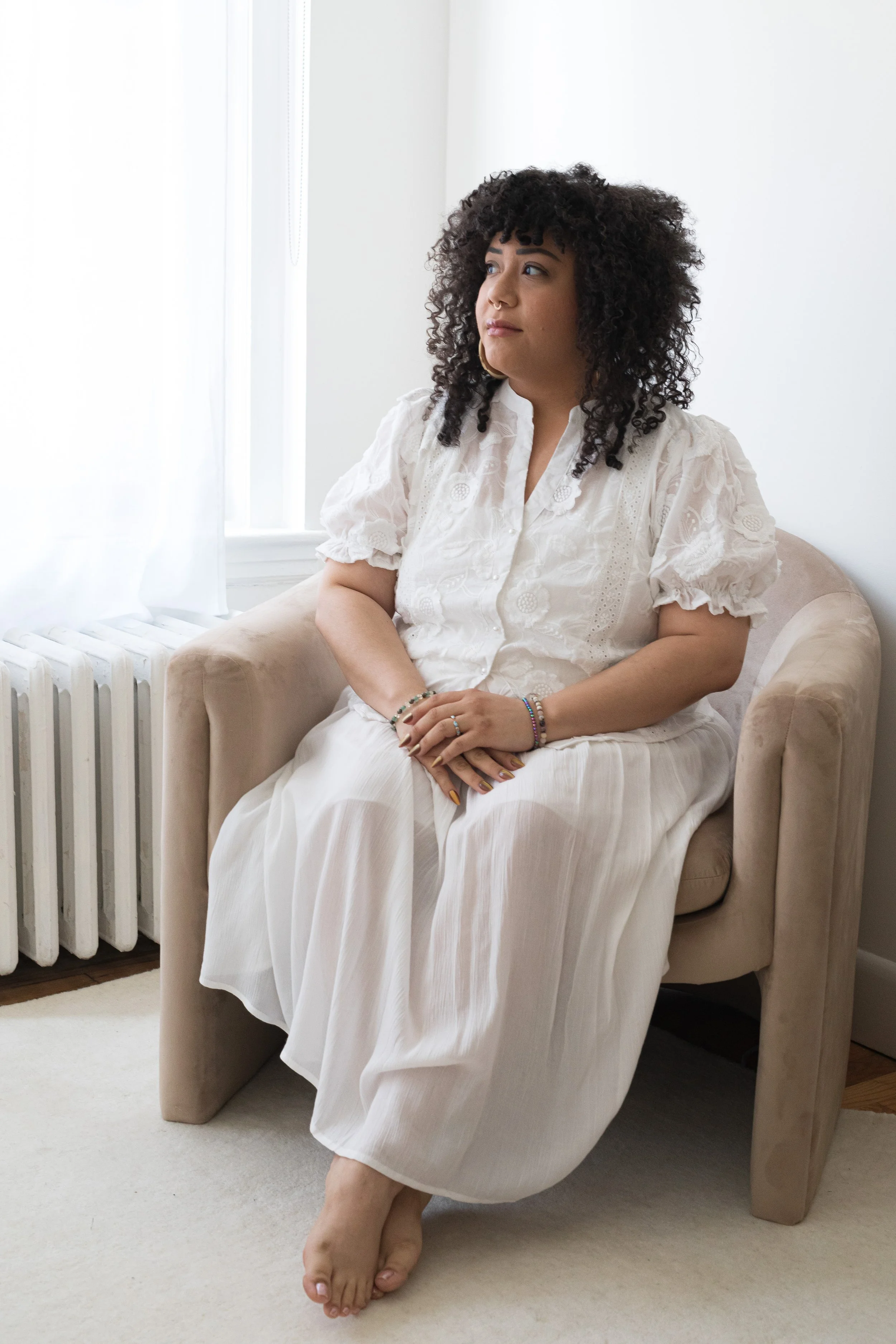 A woman with curly black hair and tan skin sitting on a beige armchair in a room with white walls and a window, wearing a white dress with sleeves, bracelets, and makeup.