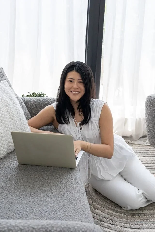 A woman with black hair in a white sleeveless top and white pants is sitting on the floor with a laptop in front of her. She is smiling and in a bright room with large windows and sheer white curtains.