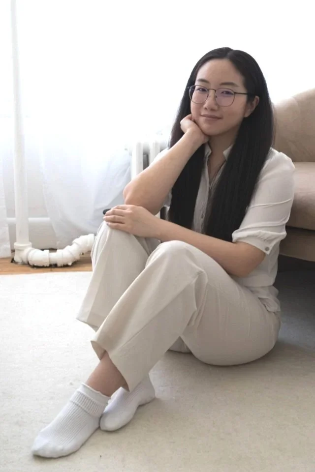 A woman with long black hair and glasses sitting on the floor, resting her head on her hand, with a relaxed expression, dressed in white pants, a white top, and white socks, in a bright room with a beige sofa and a white radiator in the background.