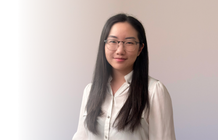 A young woman with long dark hair, glasses, and a white blouse standing against a plain light-colored wall.