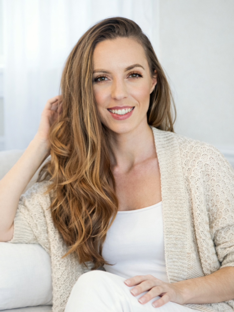 A smiling woman with long wavy red hair, wearing a white top and a beige knitted cardigan, sitting on a light-colored sofa in a well-lit room.