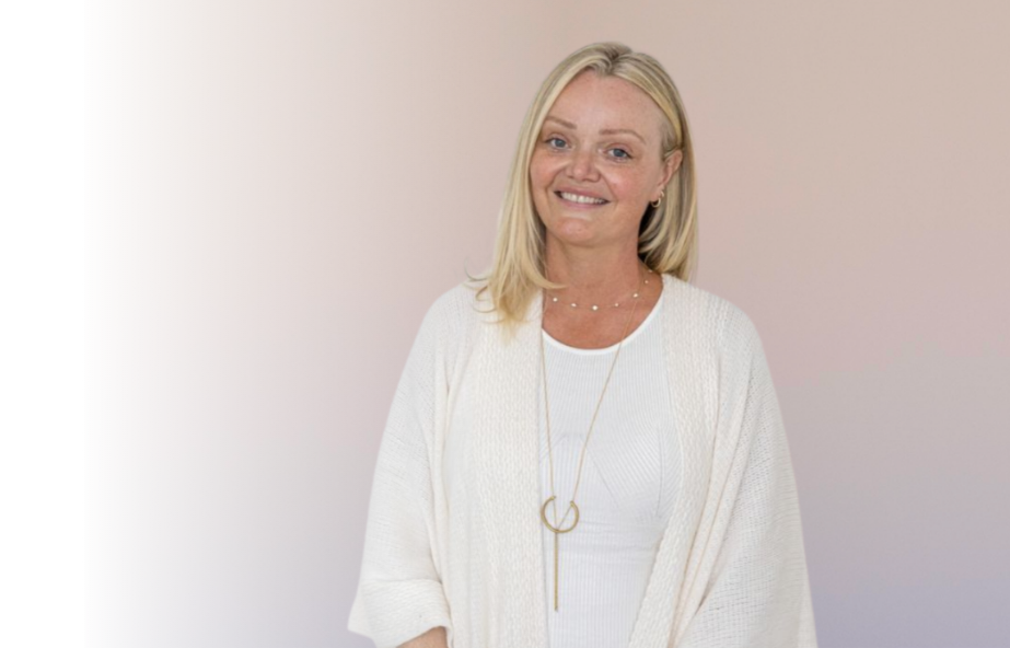 A smiling blonde woman wearing a white cardigan and necklace standing against a plain light-colored background.