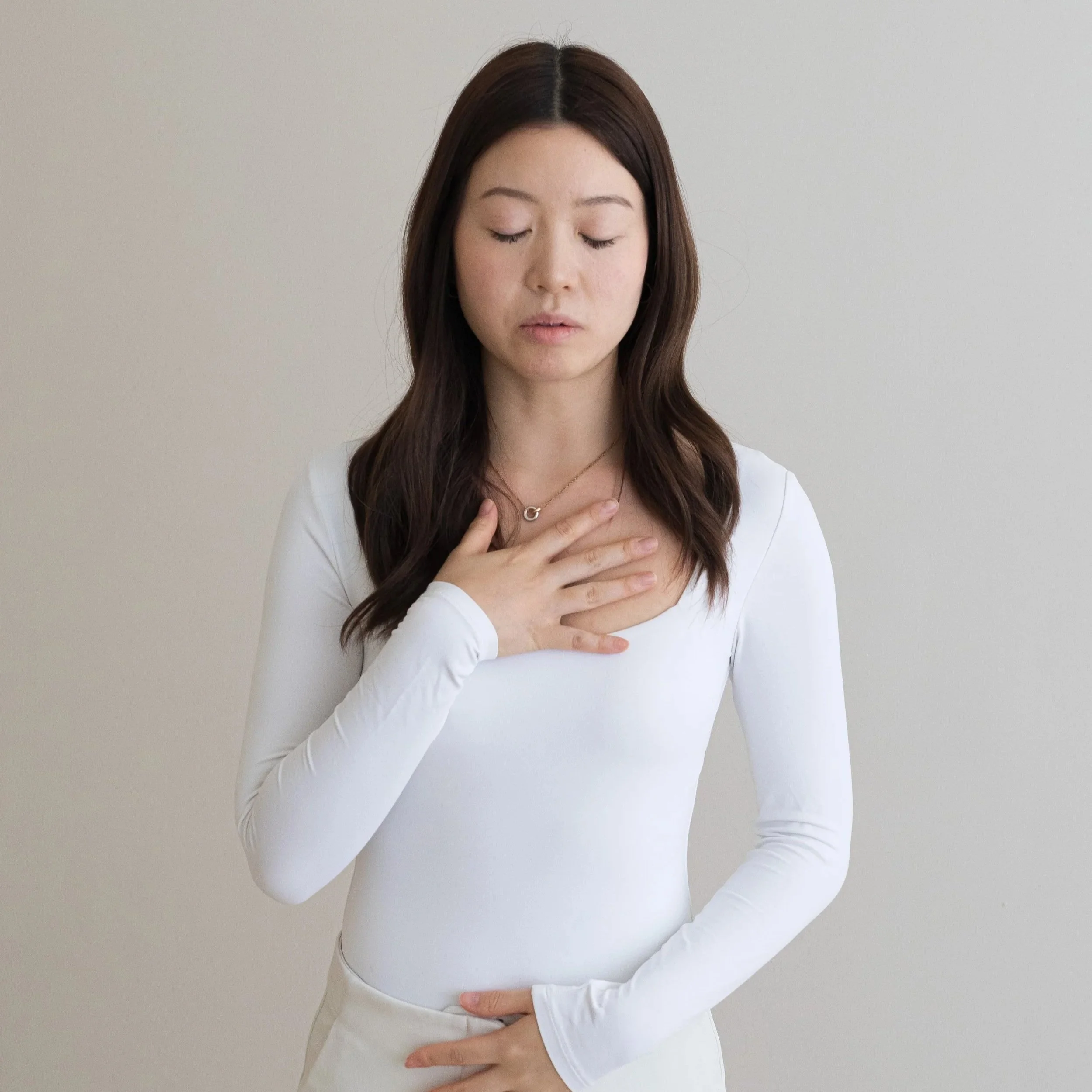 A woman with long dark hair, wearing a white long-sleeve shirt and a gold necklace, stands against a plain white background with her eyes closed, hand on chest, appearing to be in a moment of reflection or emotion.