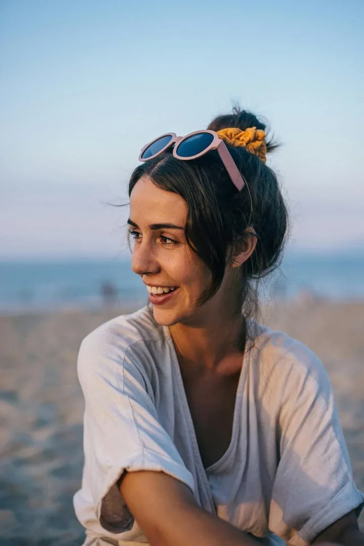 A young woman smiling and looking to the side on a beach, wearing sunglasses on her head, with a scrunchie in her hair, and a white t-shirt.