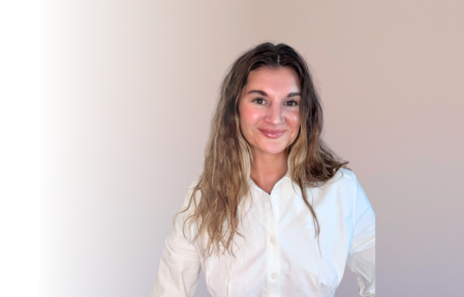 A woman with wavy, light brown hair and fair skin, wearing a white button-up shirt, smiling at the camera against a plain, light-colored background.
