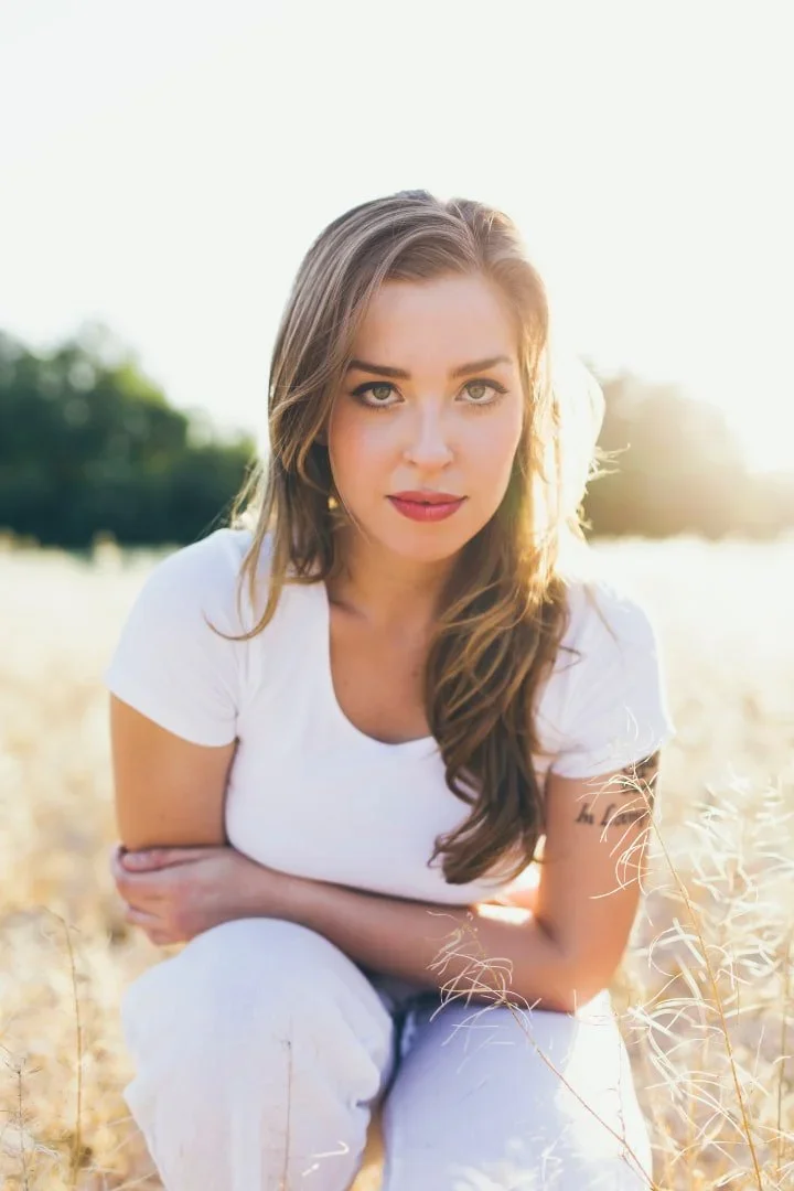 A young woman with long brown hair sitting in a field of tall grass, wearing a white t-shirt, with sunlight behind her, looking directly at the camera.