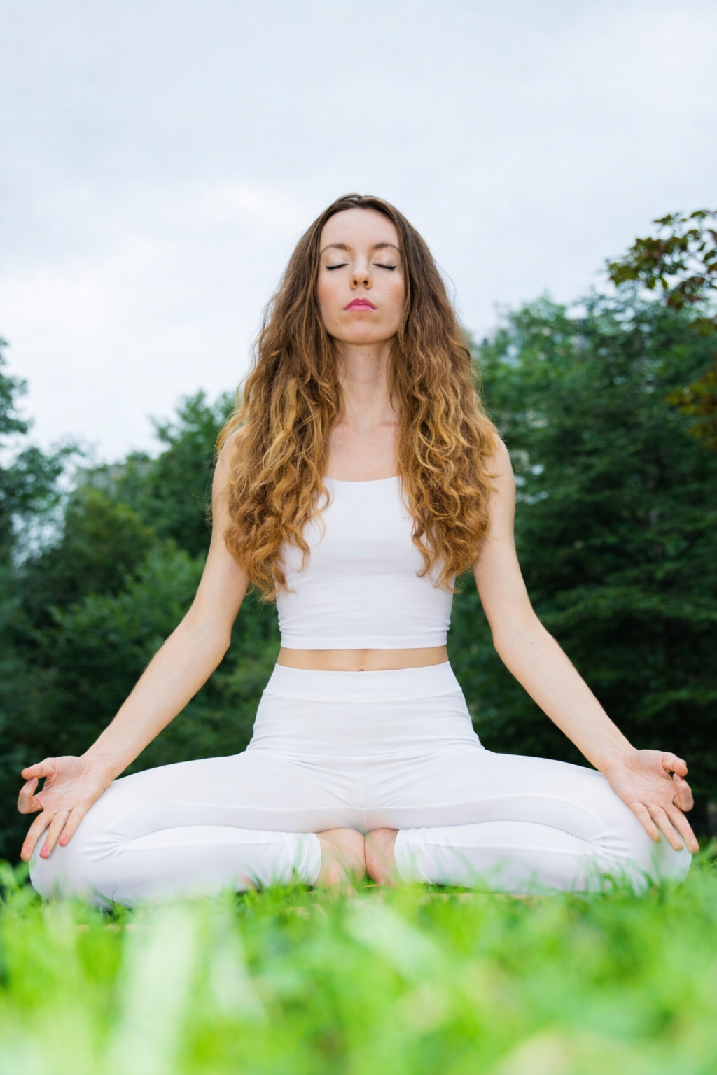 A woman with long curly hair practicing yoga outdoors in a seated meditation pose with closed eyes, wearing all white, surrounded by green trees and grass.