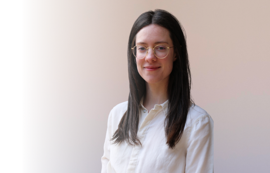 A young woman with long dark hair, wearing glasses and a white shirt, standing against a plain beige background.