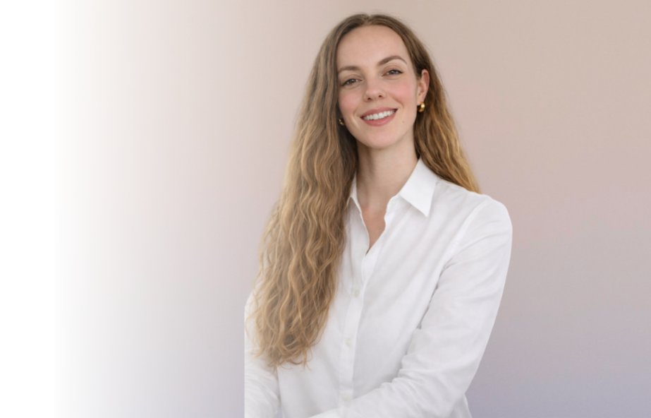 A woman with long, wavy blonde hair smiling, wearing a white button-up shirt and small gold earrings, standing against a light neutral background.