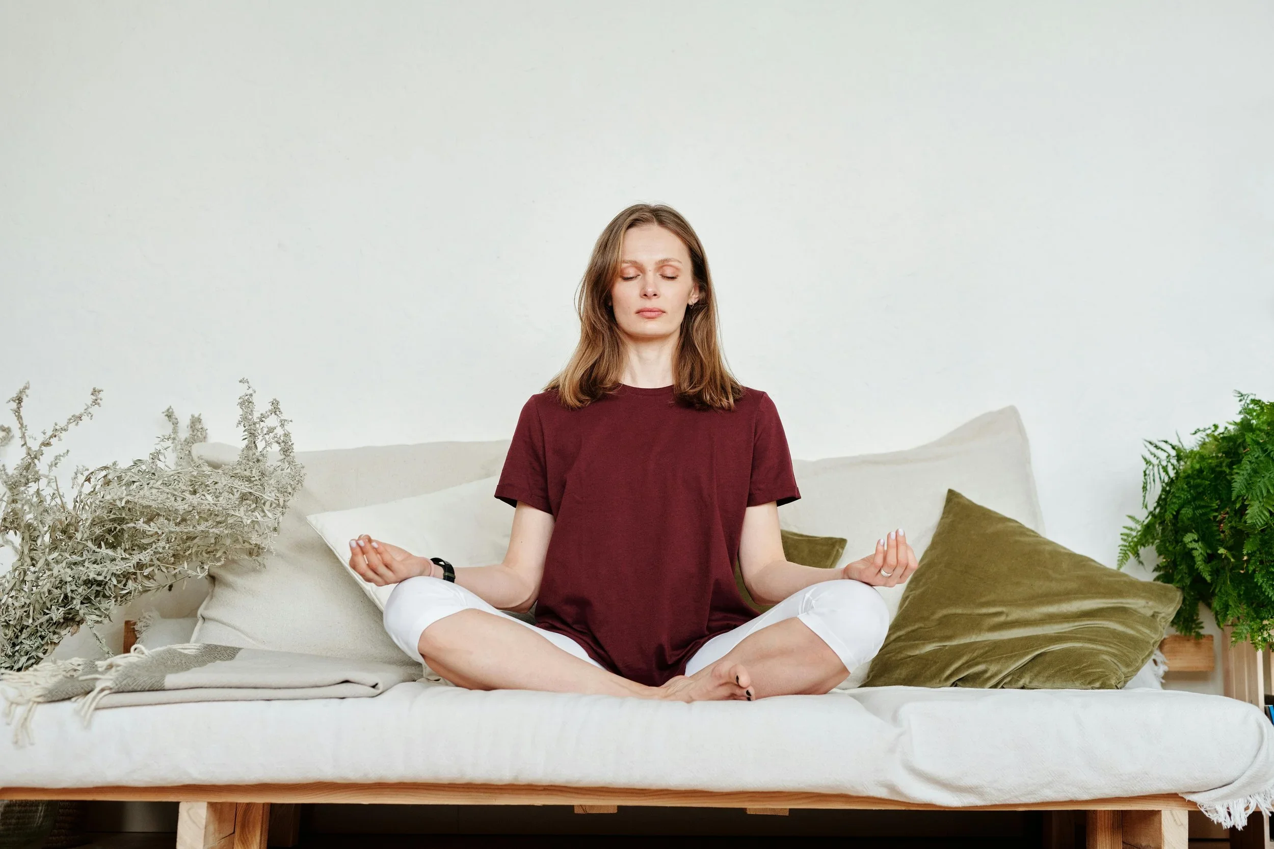 A woman practicing meditation or yoga on a bed with her legs crossed and hands resting on her knees, eyes closed in a peaceful expression, surrounded by neutral and green cushions and plants.