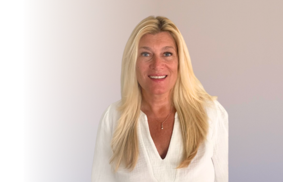 A woman with long blonde hair smiling, wearing a white top, standing against a light background.