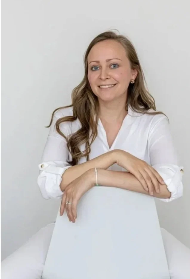 A woman with long light brown hair and blue eyes, wearing a white blouse, sitting in front of a plain white background, smiling and resting her arms on a white chair.