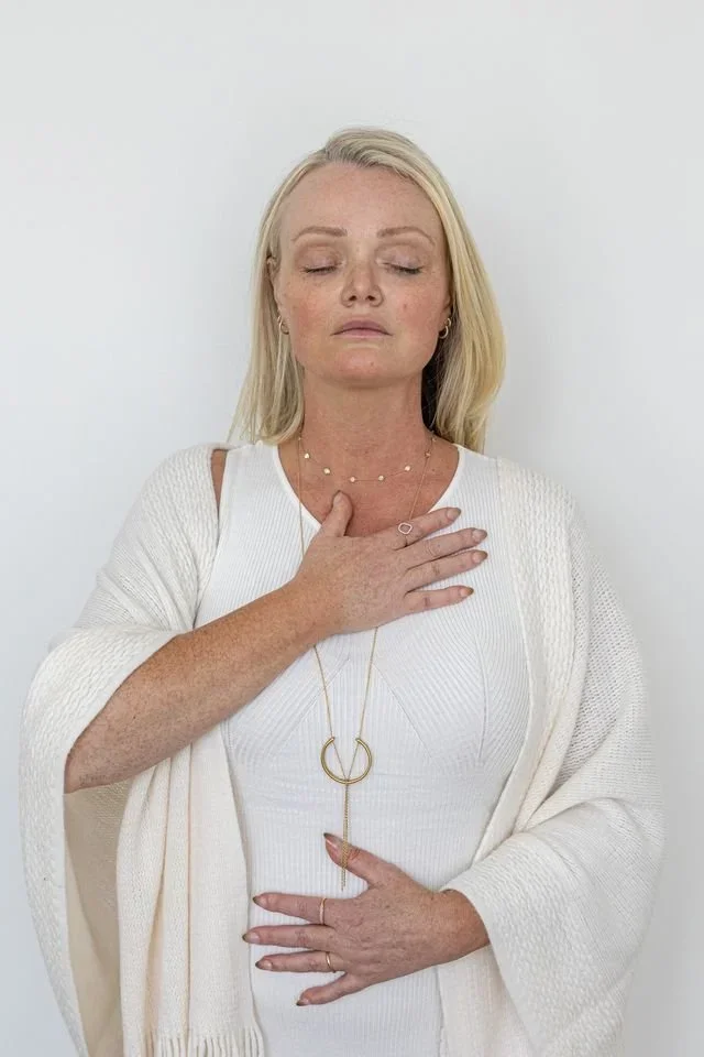 Woman with closed eyes, hand on chest, wearing white clothing and jewelry, standing against a plain white background.