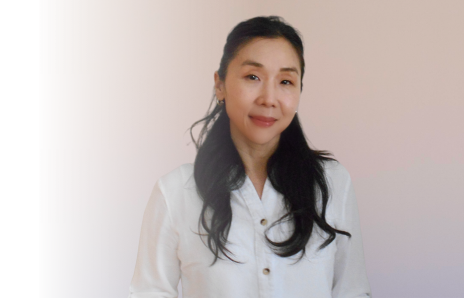 A woman with long black hair wearing a white shirt, standing against a light pink background.