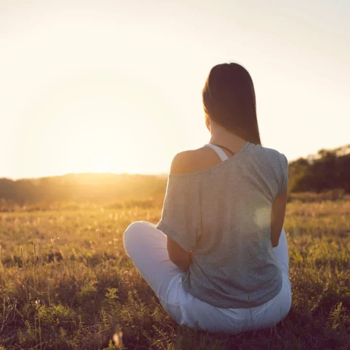 A woman sitting in a grassy field during sunset, facing away from the camera.