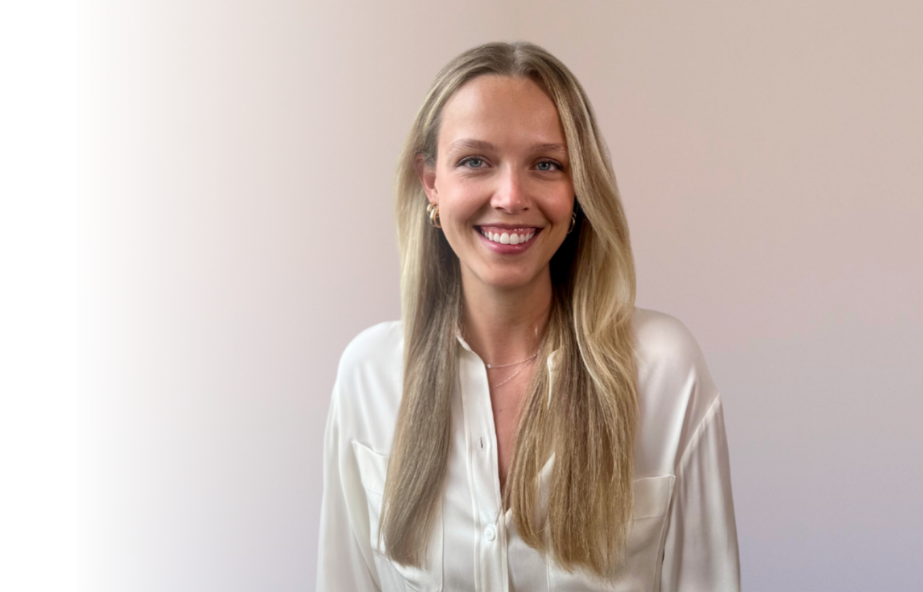 A young woman with long blonde hair smiling, wearing a white blouse, standing against a plain light background.