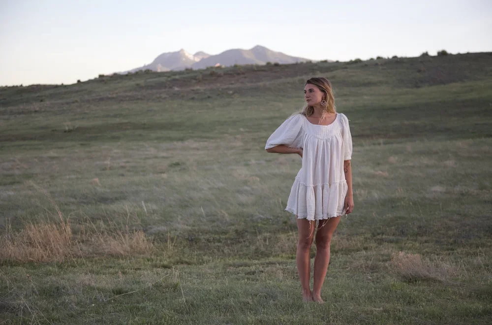 A woman in a white dress standing barefoot outdoors in a grassy field, with mountains in the background.