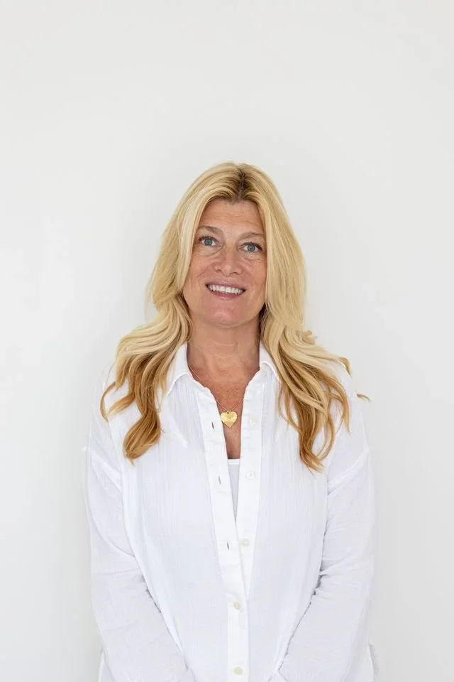 Portrait of a smiling woman with long blonde hair wearing a white shirt and a gold heart necklace against a plain white background.
