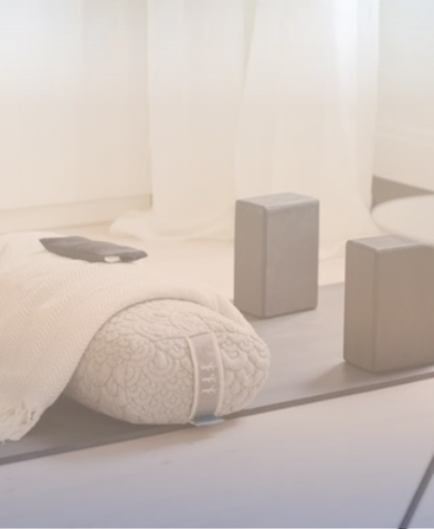 Close-up of a neatly made bed with white bedding, a rolled towel at the foot, and two gray books or boxes on a tray at the head of the bed, in a bright room with sheer curtains.