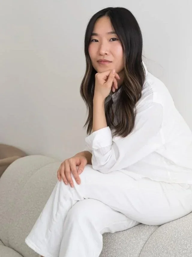 A woman with long dark hair sitting on a light-colored sofa, wearing a white outfit, resting her chin on her hand, looking at the camera.