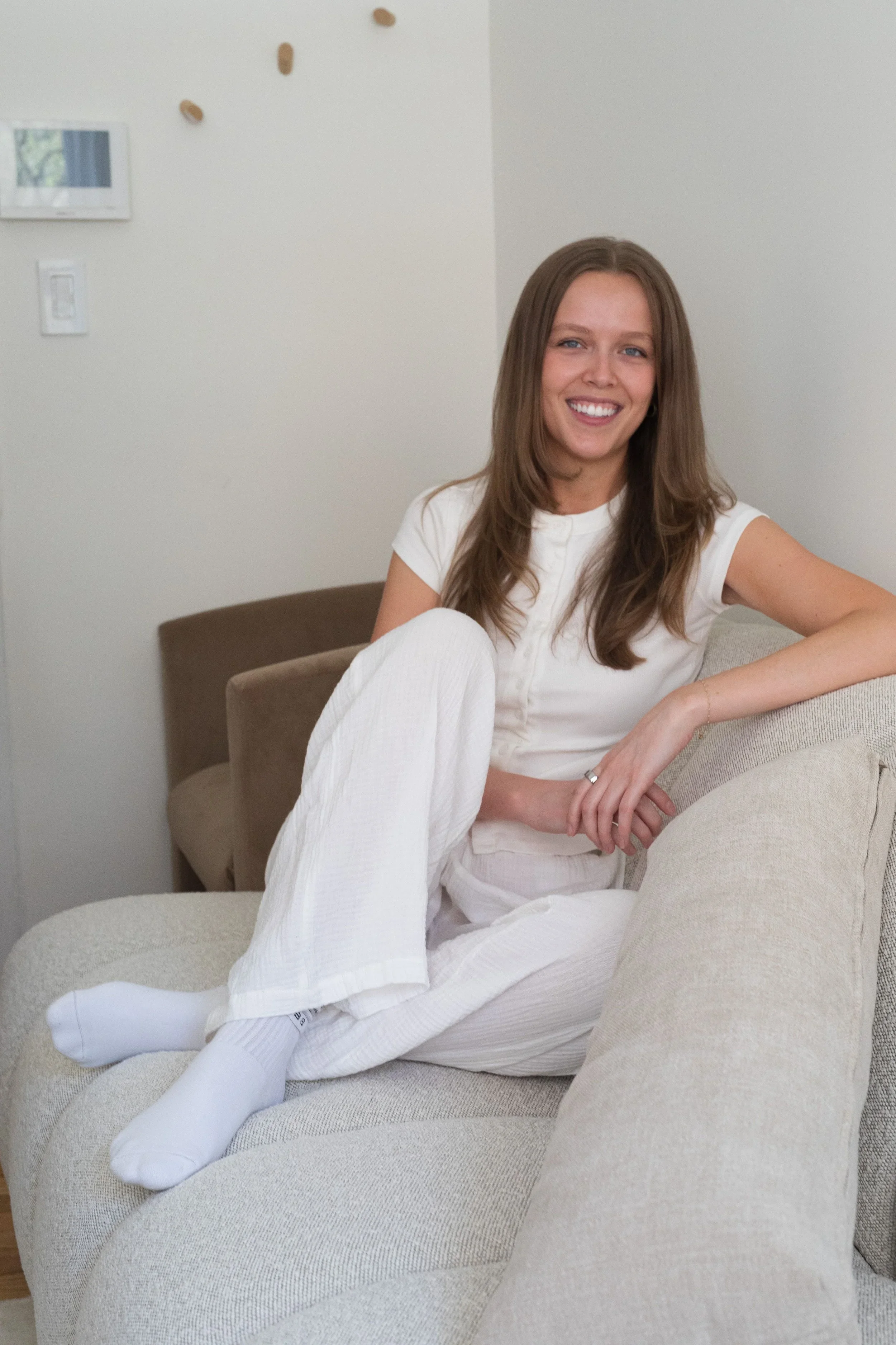 A young woman with long brown hair smiling and sitting on a beige couch in a room with white walls, wearing a white top, white pants, and white socks.