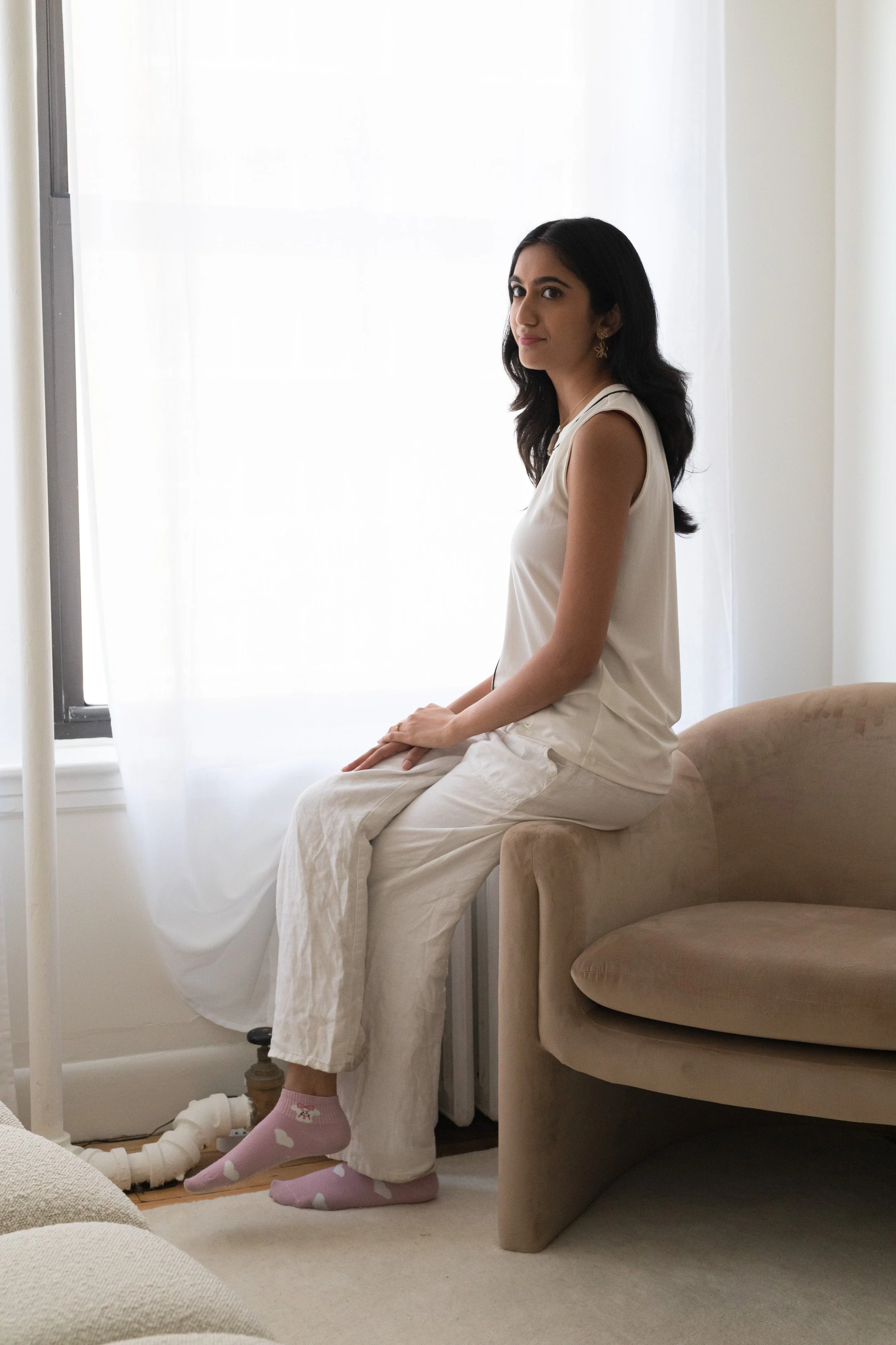A woman with dark hair sitting on a beige armchair in front of a bright window, wearing white pants, a sleeveless top, and pink socks with white hearts.
