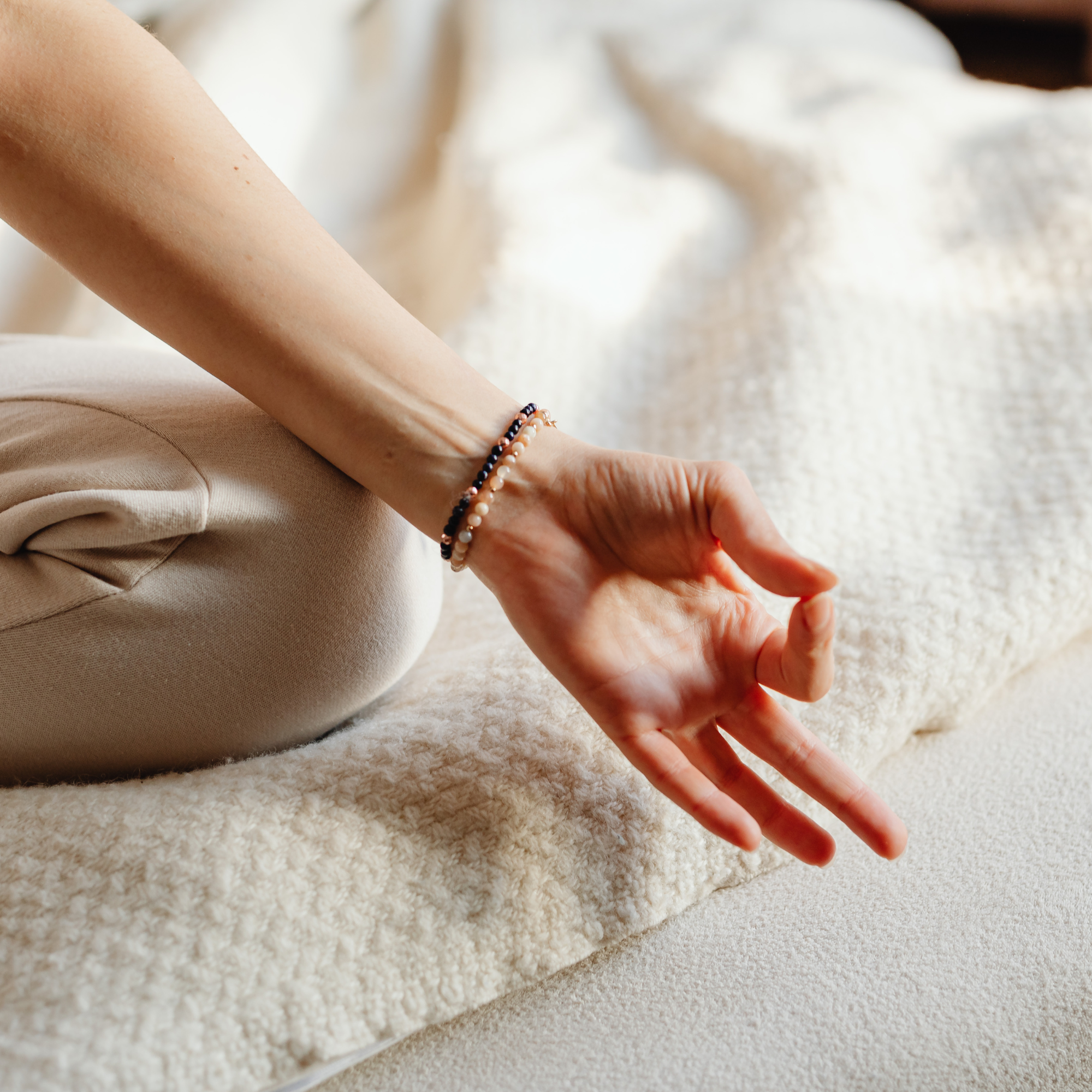 Close-up of a person's arm resting on a soft beige carpet, wearing two beaded bracelets, with a beige fabric clothing item draped nearby.